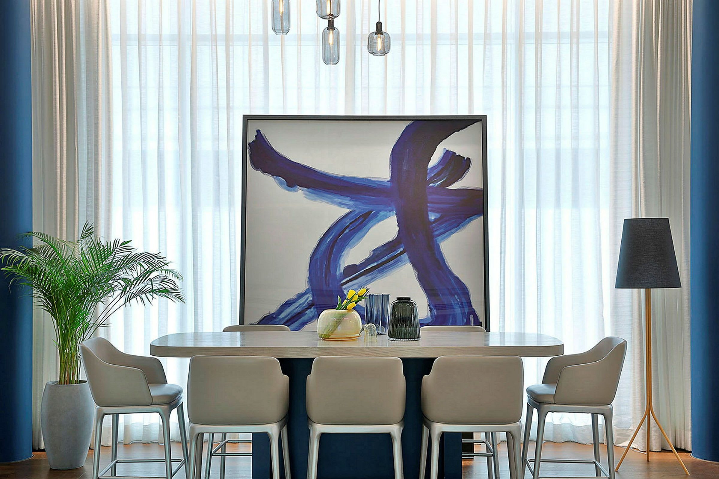 Interiör med ett modernt matbord omgiven av stolar. Ett stort konstverk med blå penseldrag står lutat mot väggen. En golvlampa och en inomhusväxt är placerade på ömse sidor om bordet. (AI-genererad text)