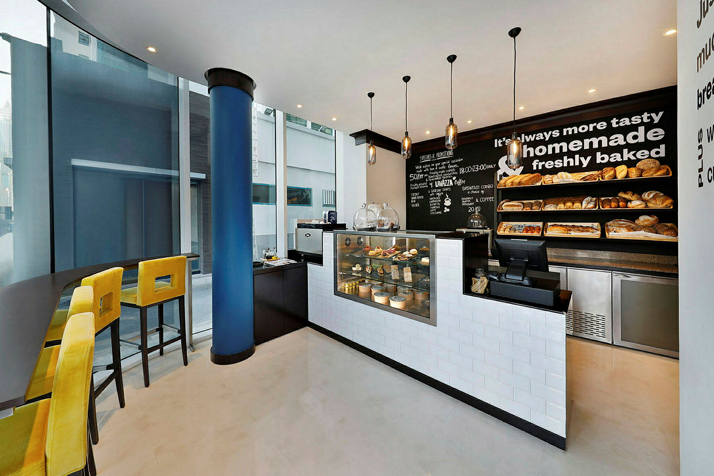 Ett kafé med modern inredning, gula barstolar vid ett högt bord vid ett stort fönster och en disk med utställda bakverk och bröd. (AI-genererad text)