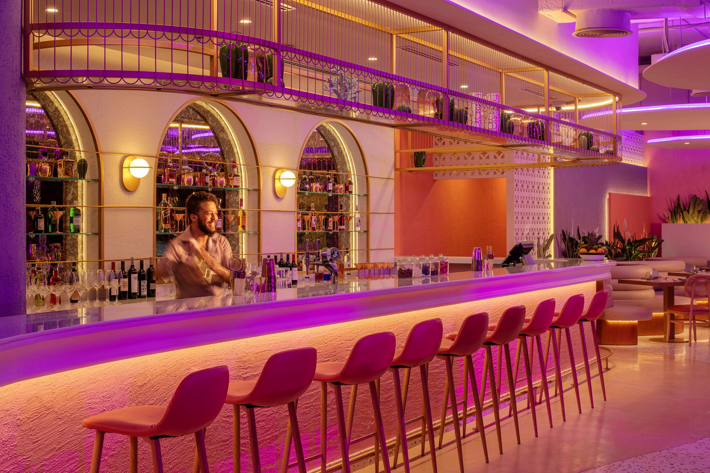 En elegant bar med rosa och lila belysning, höga stolar och flaskor på hyllor. (AI-genererad text)
