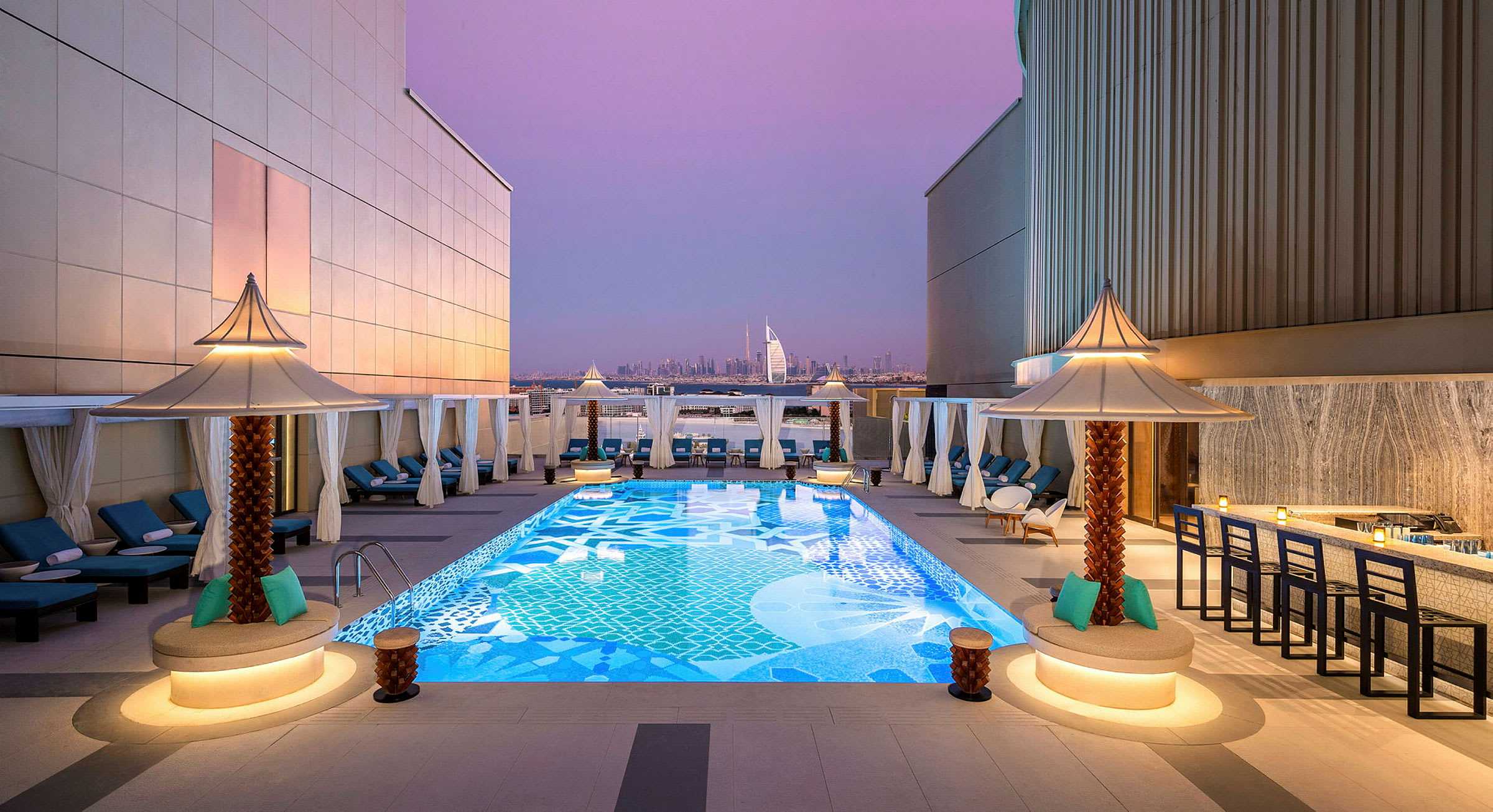 En takterrass med en dekorativ pool omgiven av solstolar och små parasoller. I bakgrunden syns Burj Al Arab och Dubais stadssilhuett under en lila kvällshimmel. (AI-genererad text)