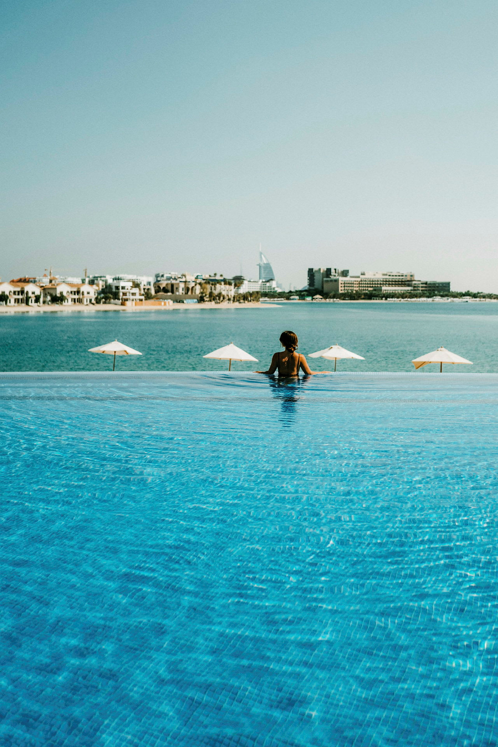 En person står i en infinitypool och tittar ut mot havet och staden i bakgrunden. (AI-genererad text)