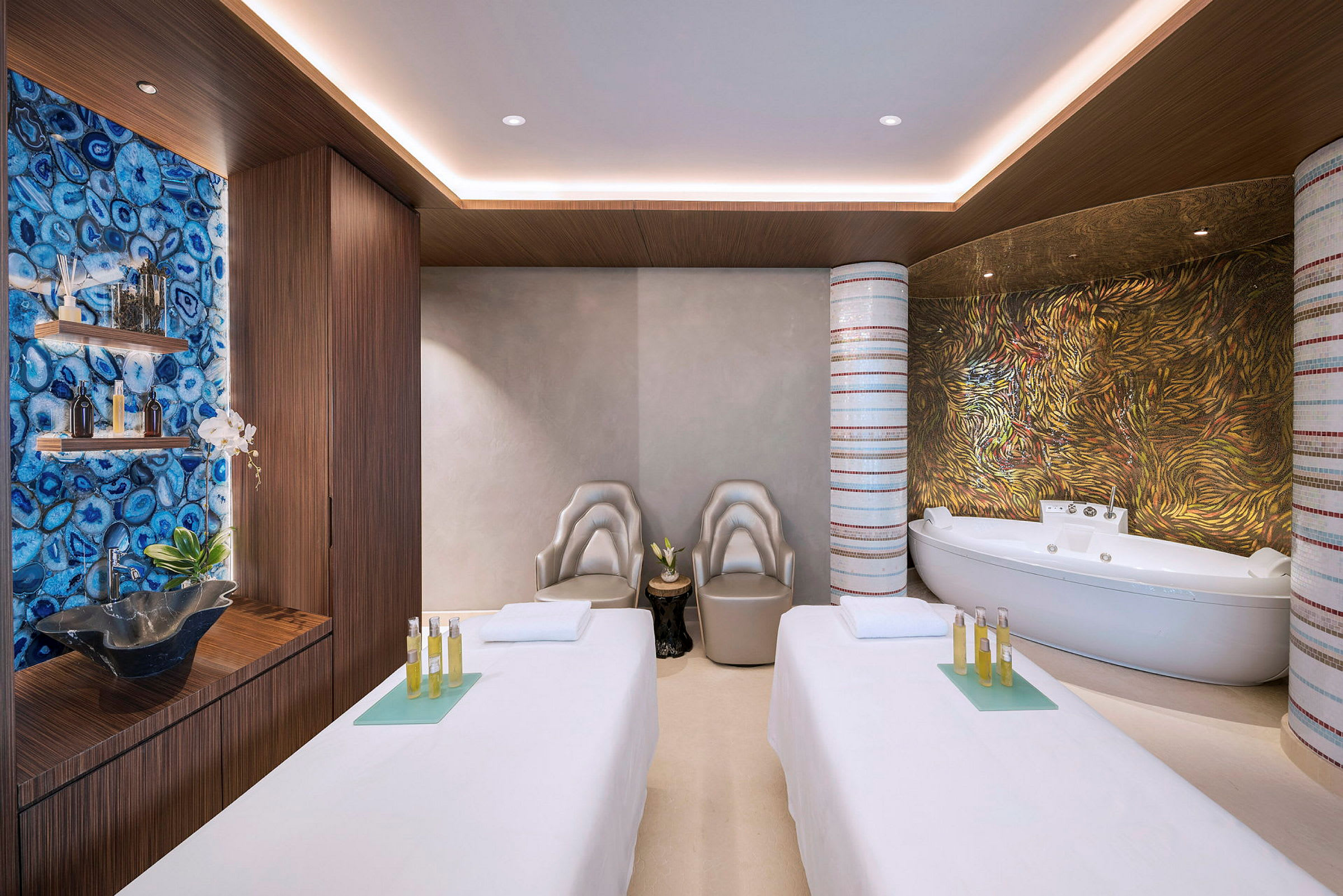 Ett spa-rum med två massagebänkar, moderna stolar och ett badkar. Bakgrunden har färgglada mönstrade väggar och hyllor med dekorationer. (AI-genererad text)