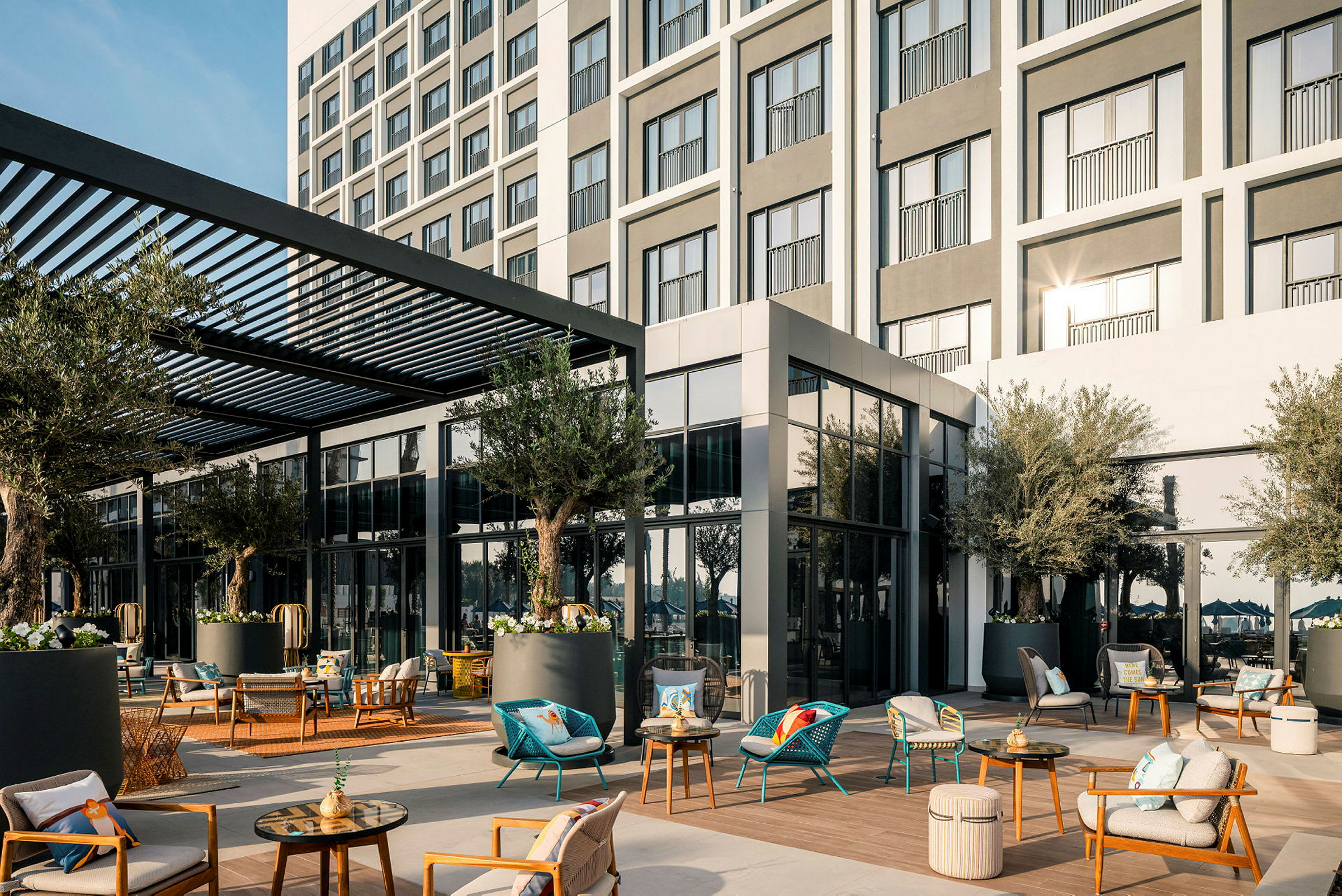 Hotellfasad med möblerad uteplats, skuggad av en pergola, omgiven av växter. (AI-genererad text)
