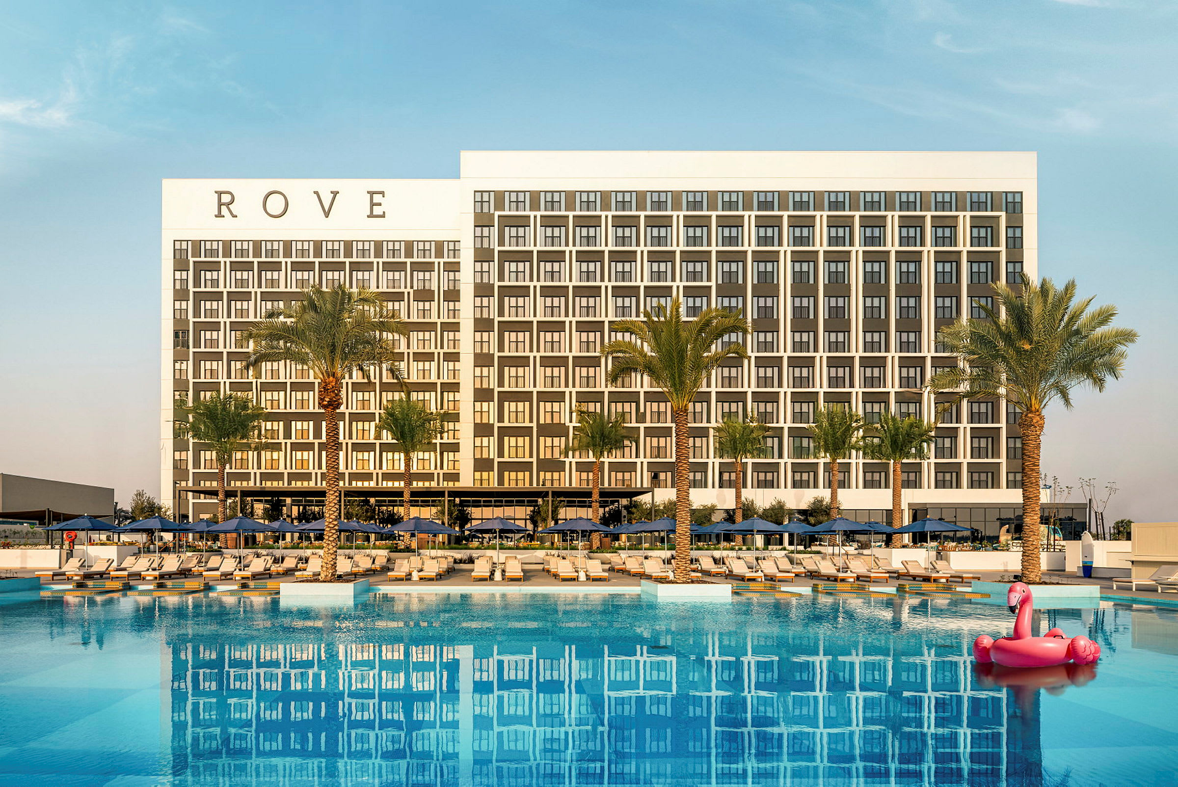 Bild av ett modernt hotell med en stor pool och palmer i förgrunden. En rosa flamingo-flytande leksak syns i poolen. (AI-genererad text)