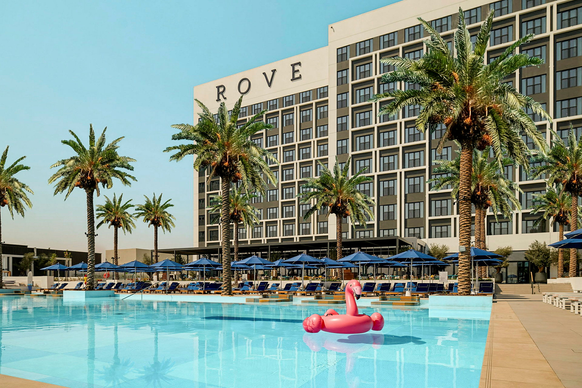 En byggnad märkt 'ROVE' bakom en pool med palmer och solstolar runtom samt en rosa flamingo uppblåsbar leksak i vattnet. (AI-genererad text)