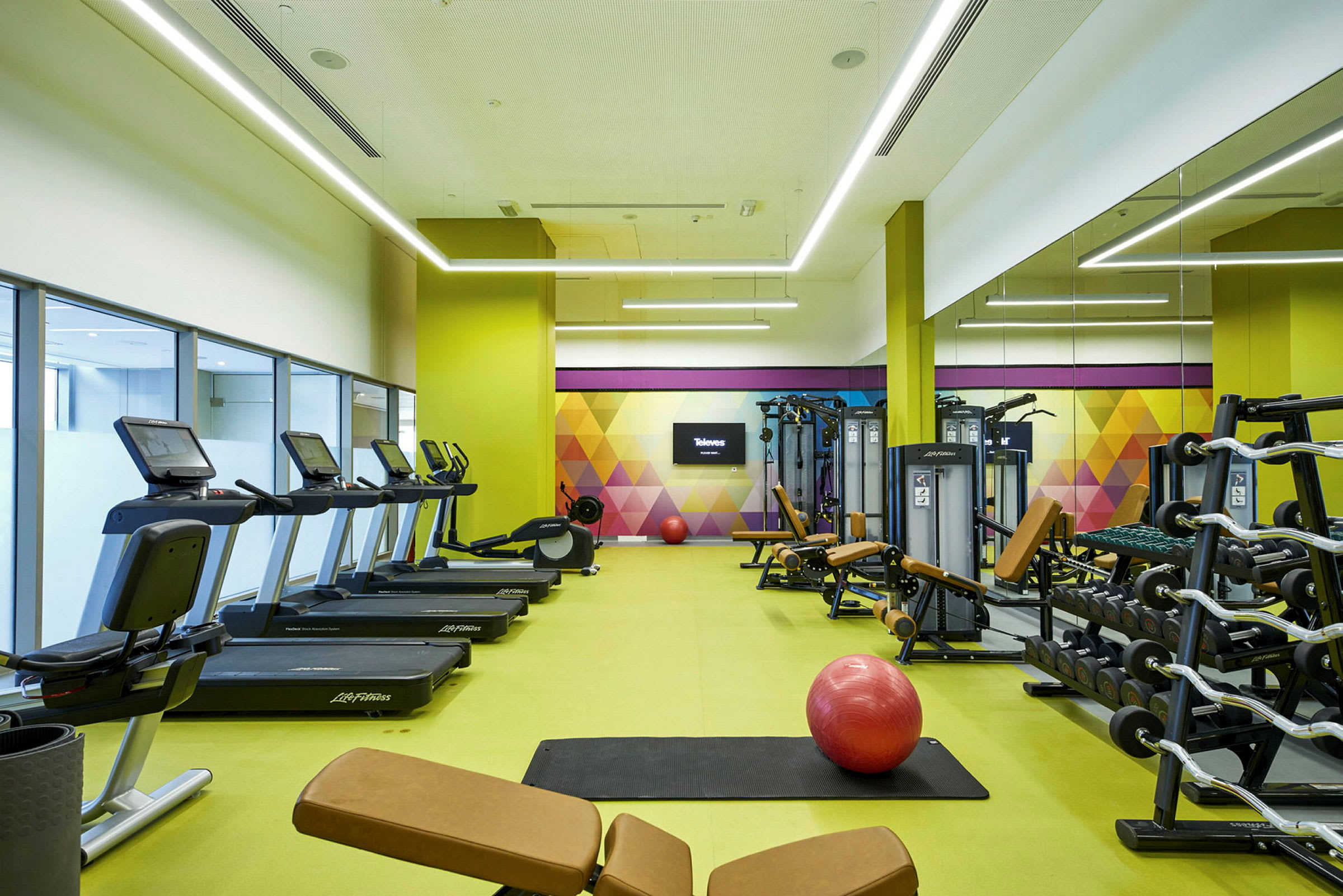Ett gym med träningsmaskiner, löpband och hantlar, samt en stor röd pilatesboll på en matta. (AI-genererad text)