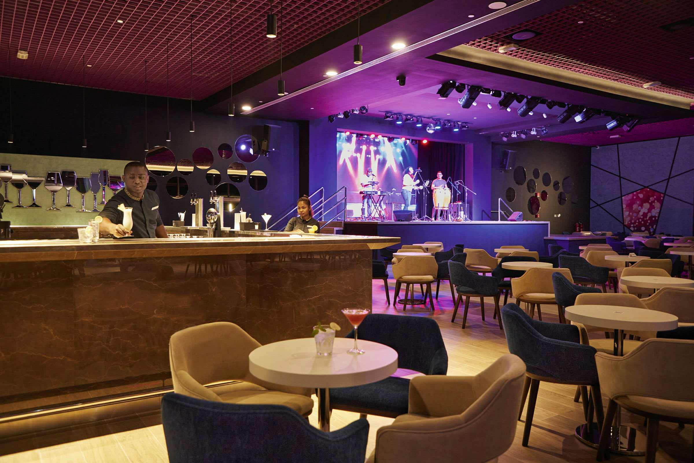 En elegant lounge med sittplatser runt små bord, en bardisk i förgrunden och en scen med musiker i bakgrunden. (AI-genererad text)