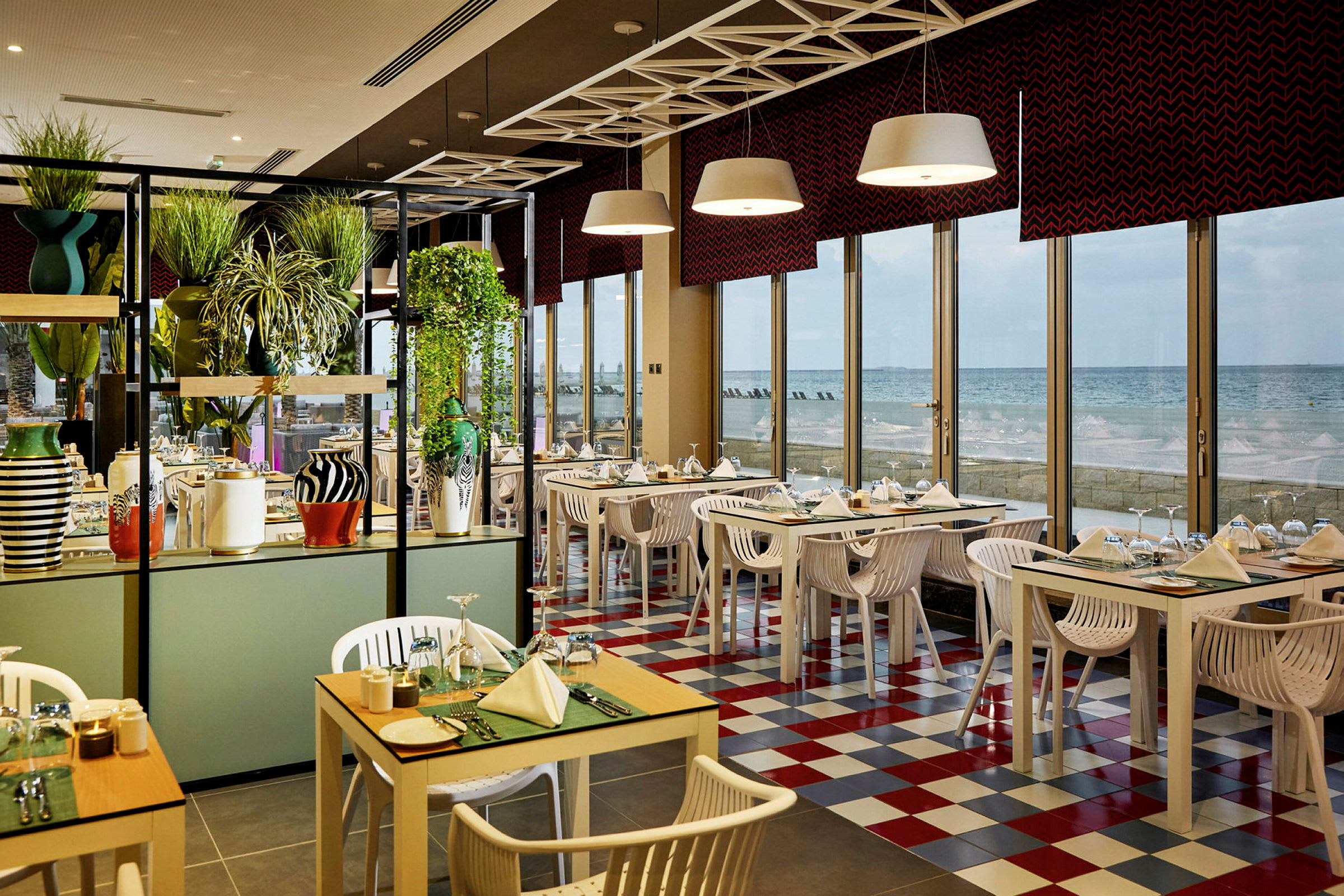 En restaurang med färgglada bord och stolar, stora fönster och utsikt över havet. (AI-genererad text)