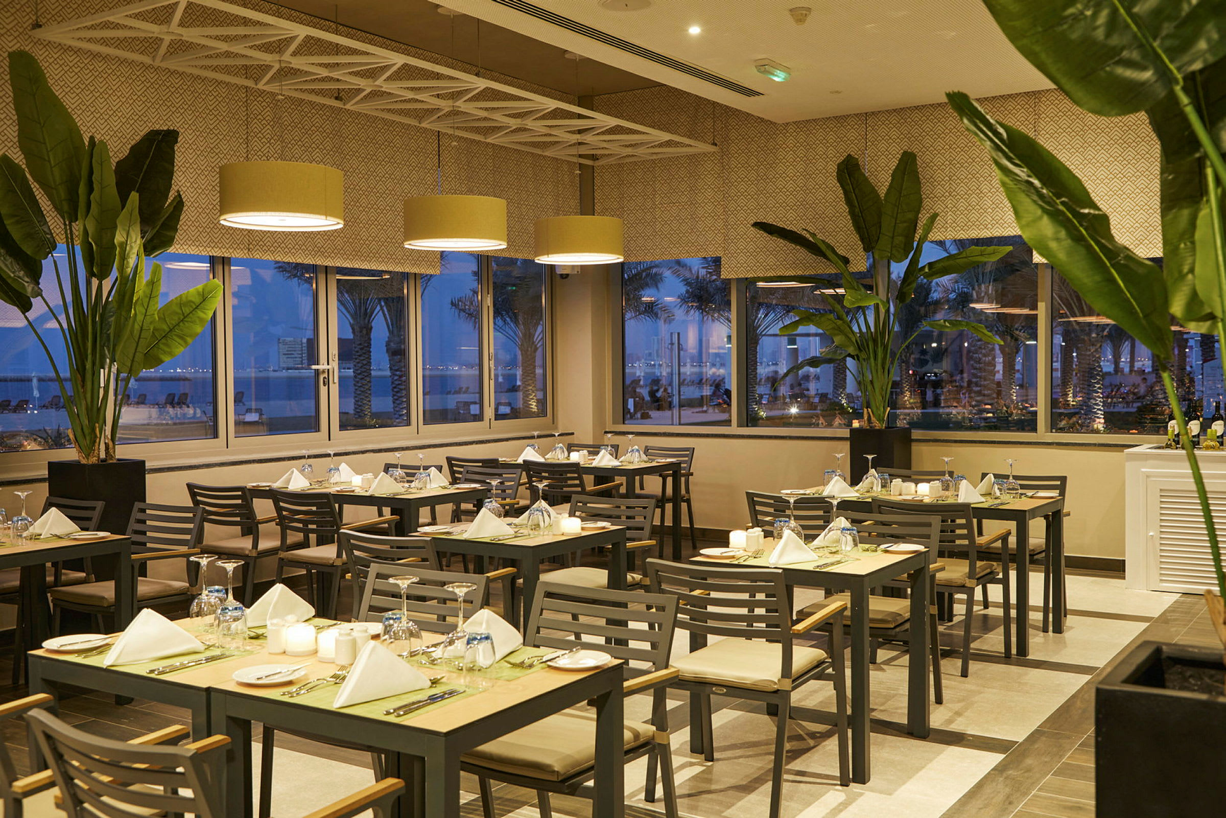 Restaurangmiljö med dukade bord, gröna växter och panoramafönster med kvällsutsikt. (AI-genererad text)