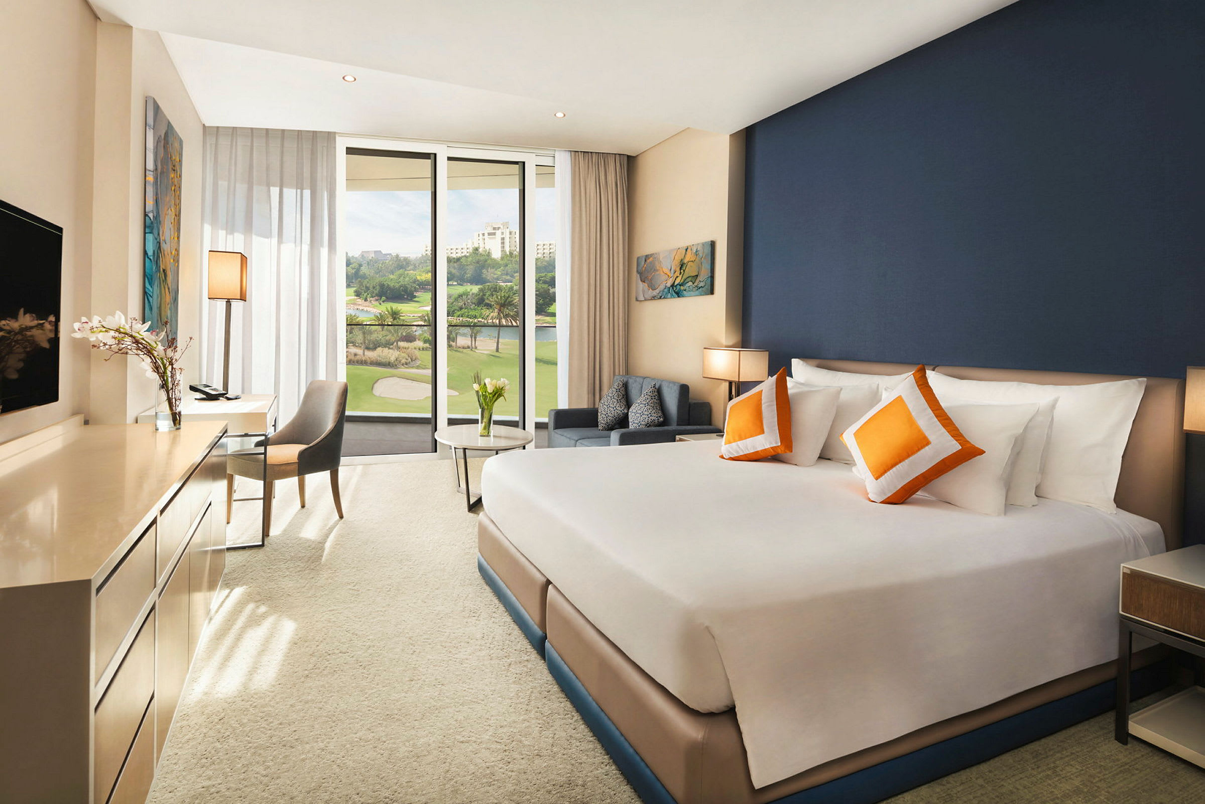 Ett välbelyst hotellrum med en stor säng som har vita sängkläder och orange prydnadskuddar. Rummet har en modern inredning, en skrivbordshörna och ett stort fönster med utsikt över gröna träd och byggnader. (AI-genererad text)