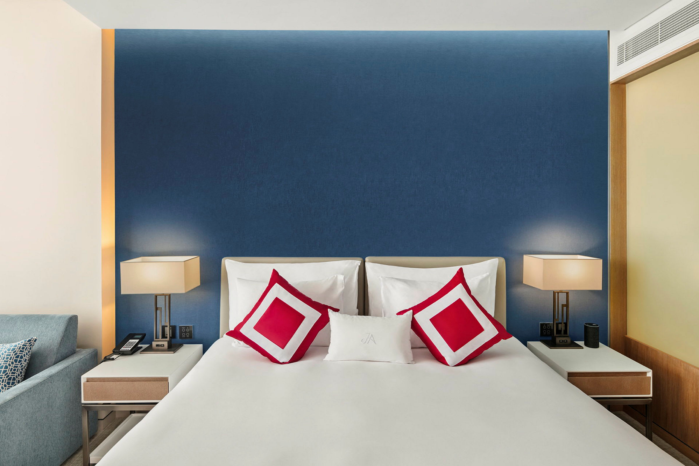 Ett modernt, smakfullt hotellrum med en dubbelsäng som har vit sängklädsel och röda dekorativa kuddar. På vardera sidan om sängen finns vita, stilrena sängbord med bordslampor. (AI-genererad text)