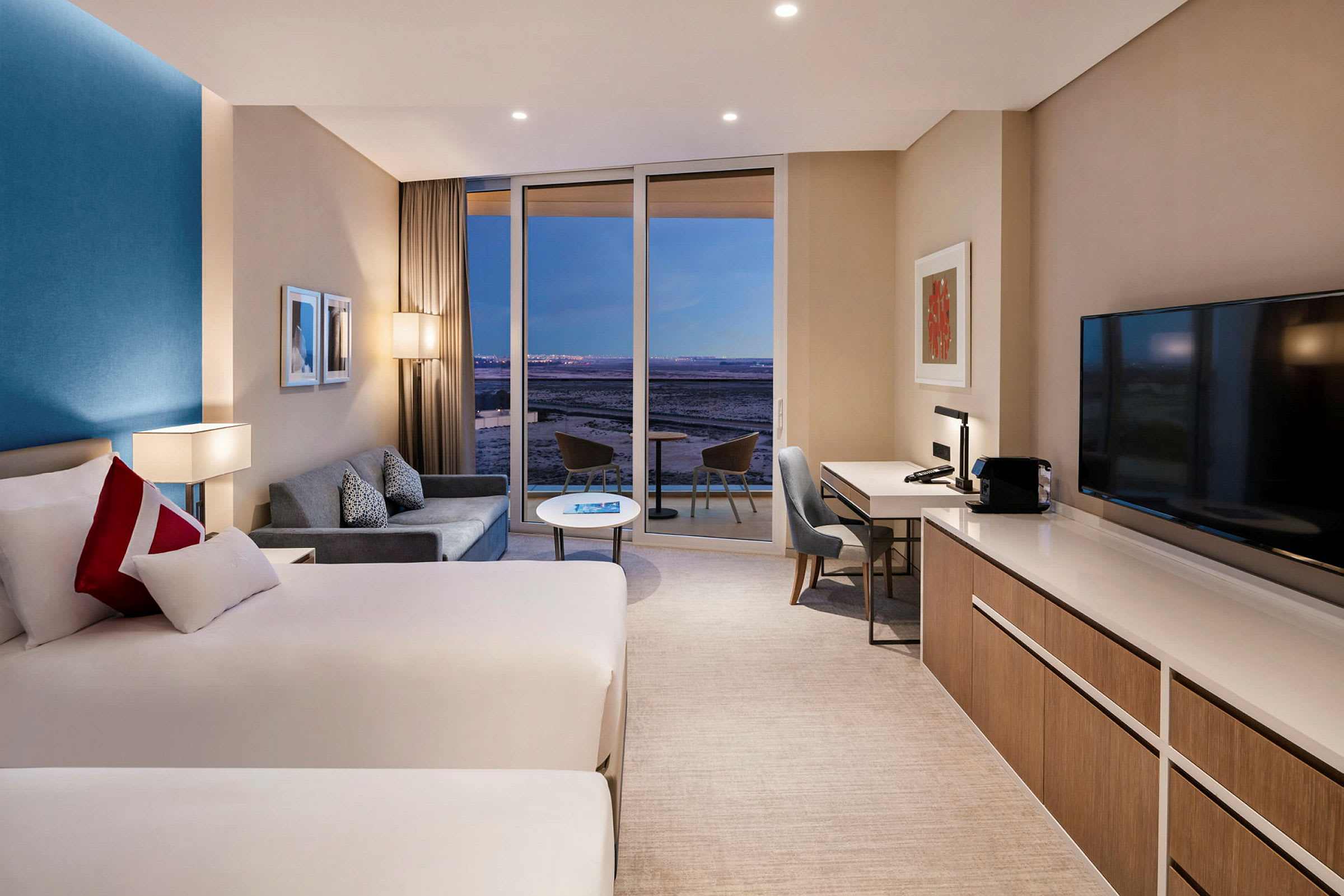 Ett modernt hotellrum med två sängar, en soffa, skrivbord och en stor glasdörr som leder ut till en balkong. (AI-genererad text)