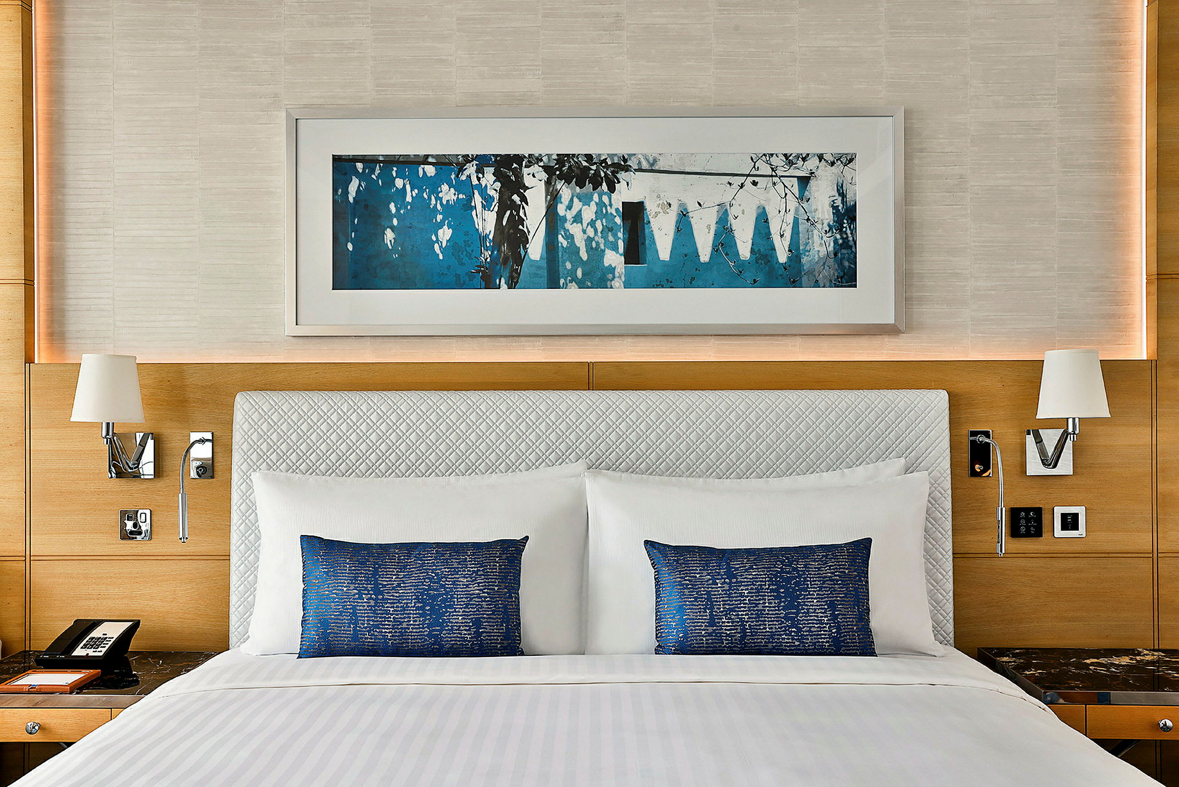 Bild på en hotellsäng med vita sängkläder och blå kuddar, omgiven av en inredning med träpaneler och en väggkonst ovanför. (AI-genererad text)