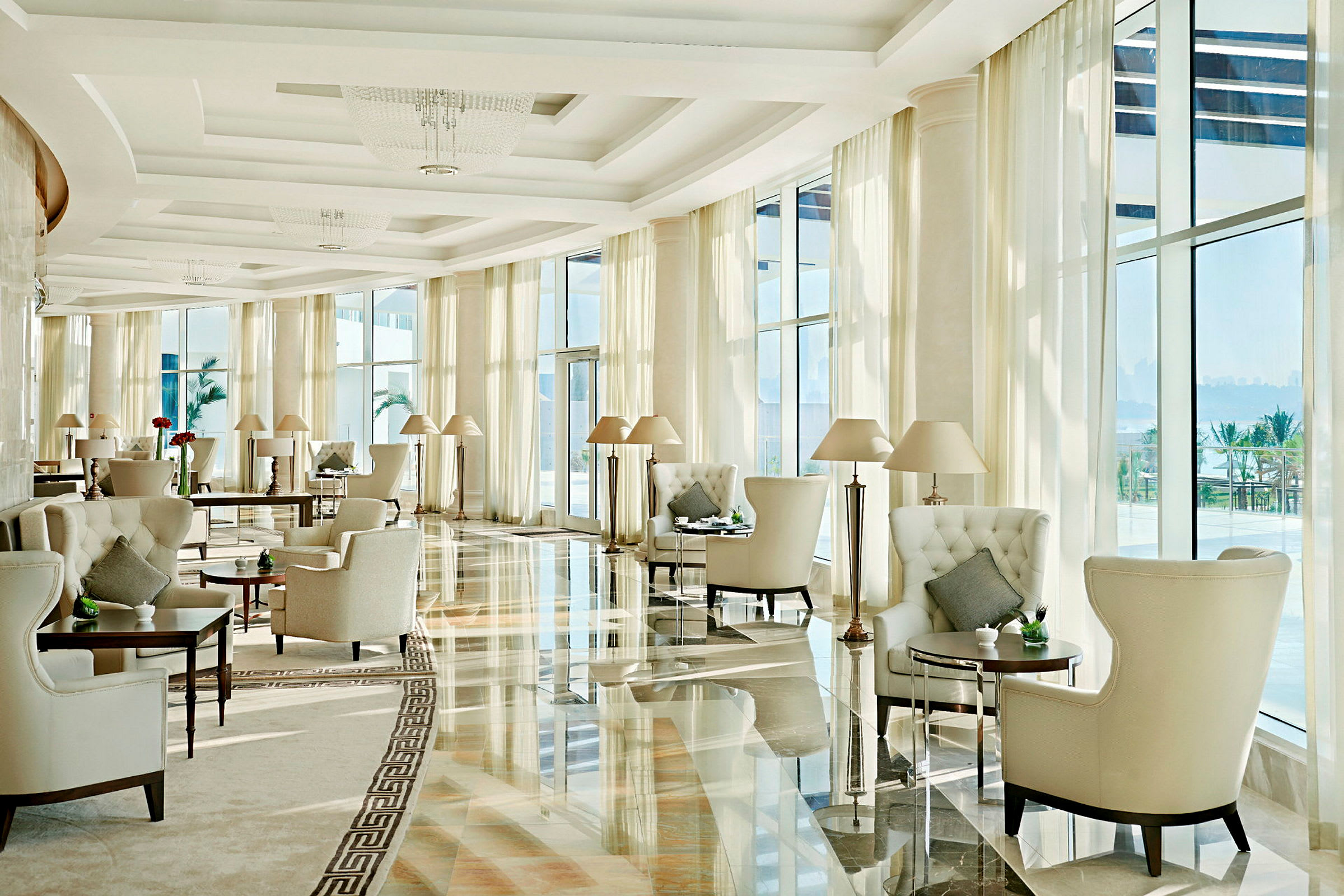 Ett ljust och elegant möblerat hotellrum med stora fönster, bekväma fåtöljer och små bord. Utsikt över palmer och hav syns i bakgrunden. (AI-genererad text)