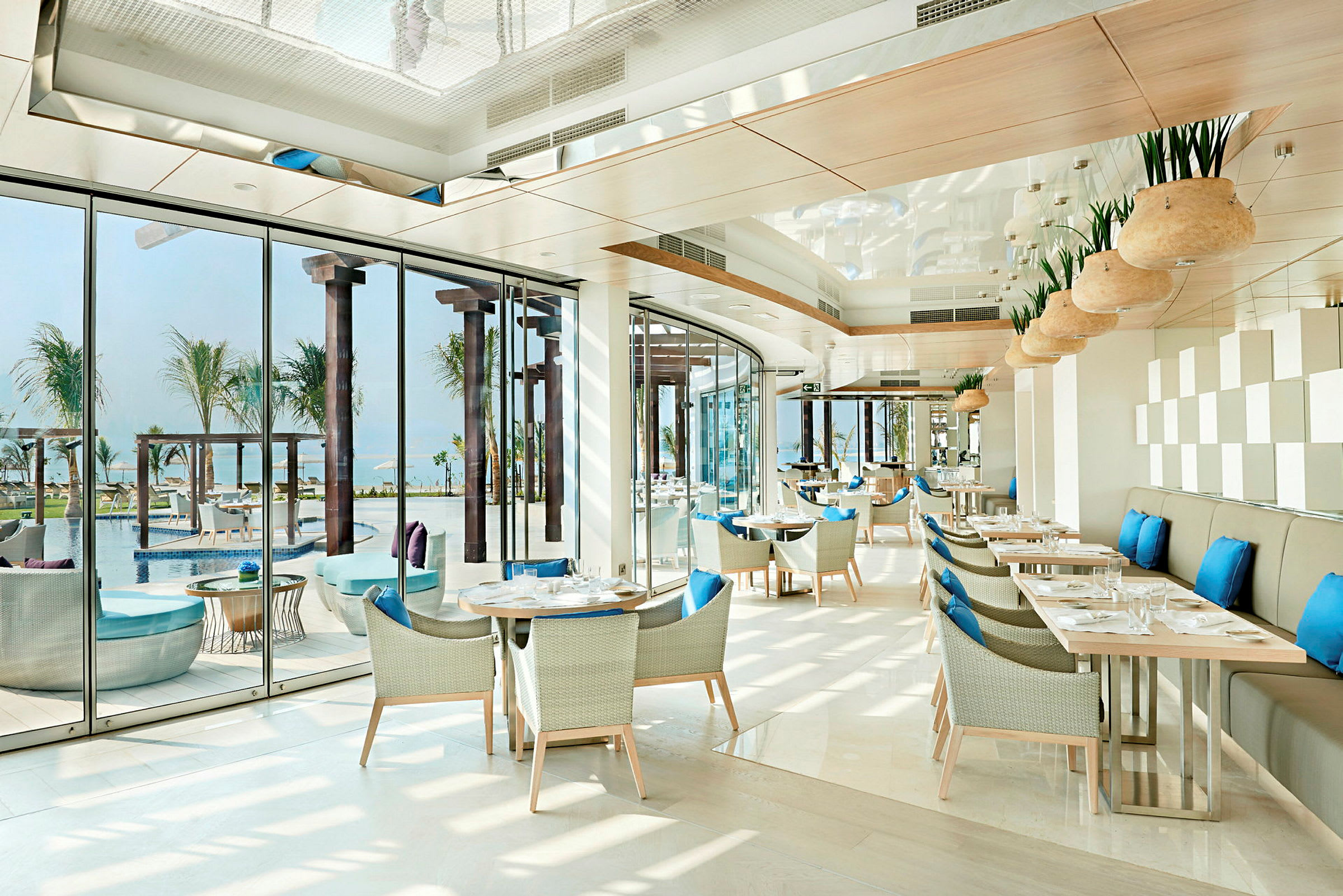 En ljus och modern restaurang med blåa kuddar på stolar och utsikt över en pool och palmer. (AI-genererad text)
