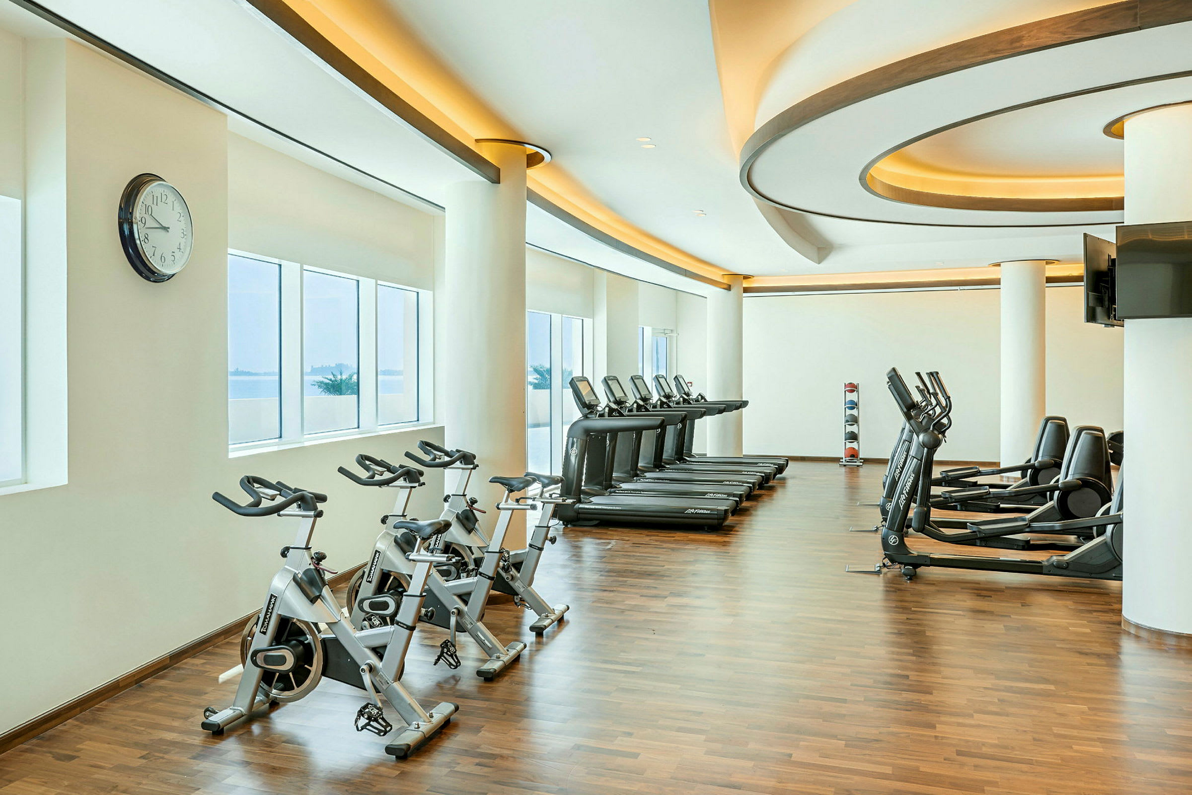 Ett gym med träningsmaskiner inklusive cyklar, löpband och andra fitnessmaskiner. Rummet har stora fönster som släpper in naturligt ljus. (AI-genererad text)