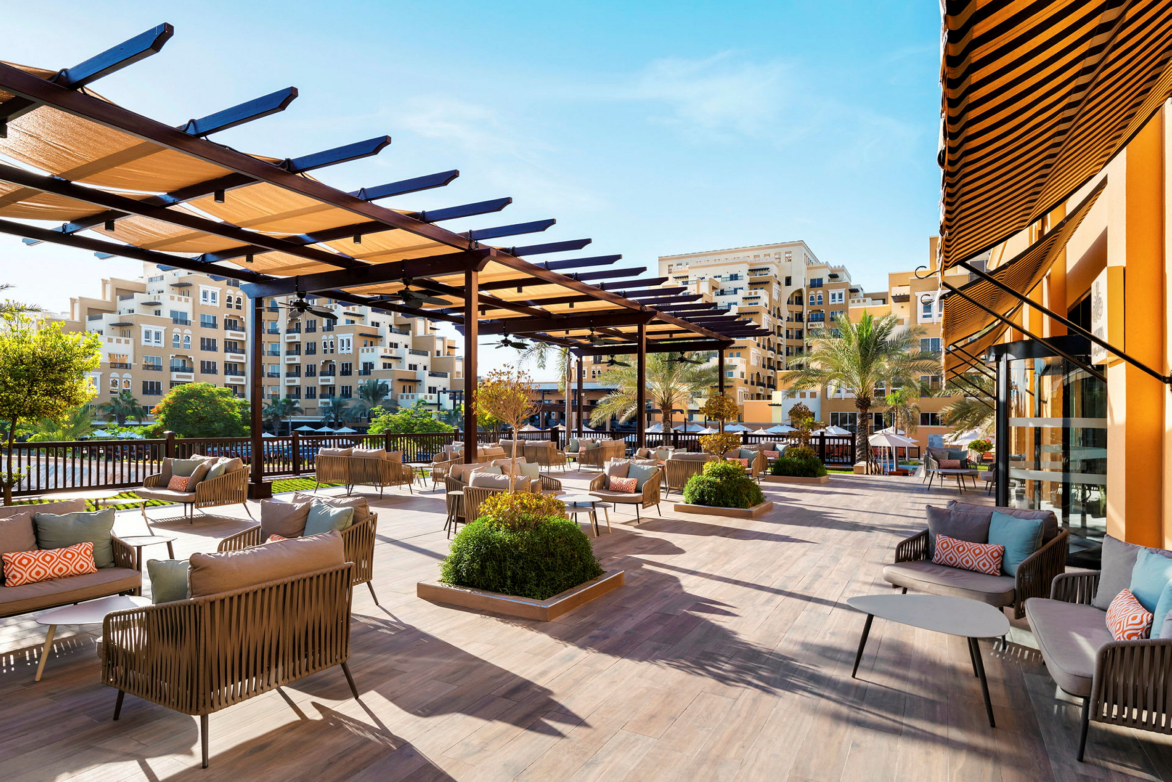 En utomhusterrass med moderna sittgrupper och en pergola under en klarblå himmel. I bakgrunden ses byggnader och palmer. (AI-genererad text)