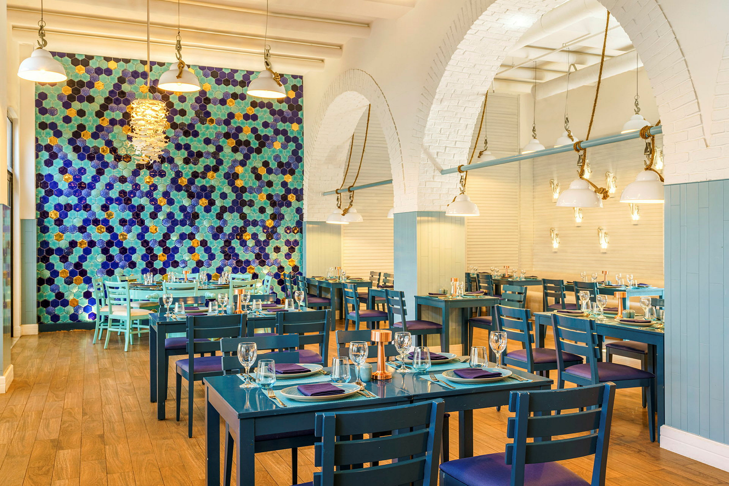 Bild på en restaurang med blå bord och stolar, en vägg med färgglada hexagonala mönster, och taklampor i modern design. (AI-genererad text)