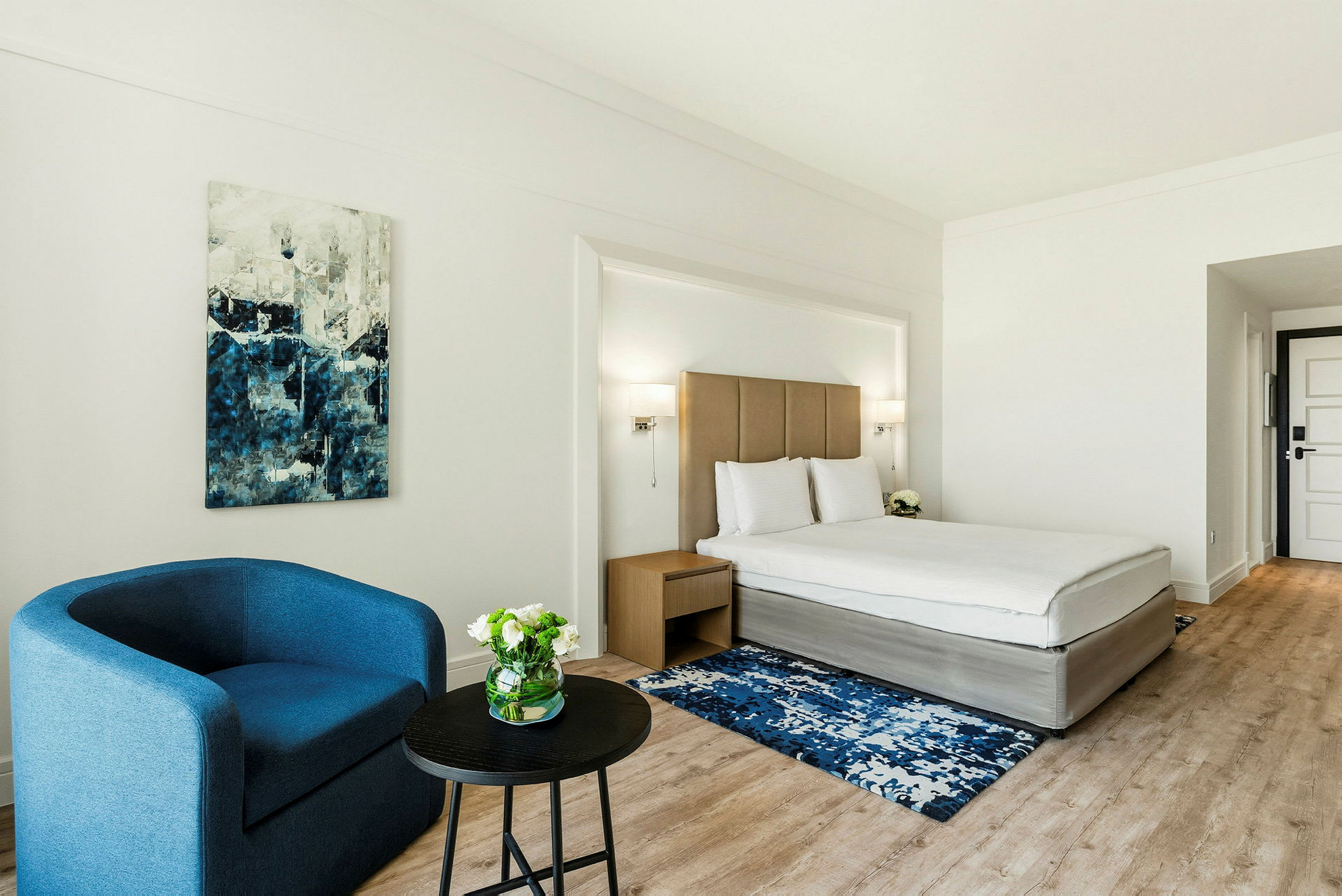 Ett hotellrum med en bäddad säng, ett blått fåtölj, ett soffbord och en målning på väggen. (AI-genererad text)