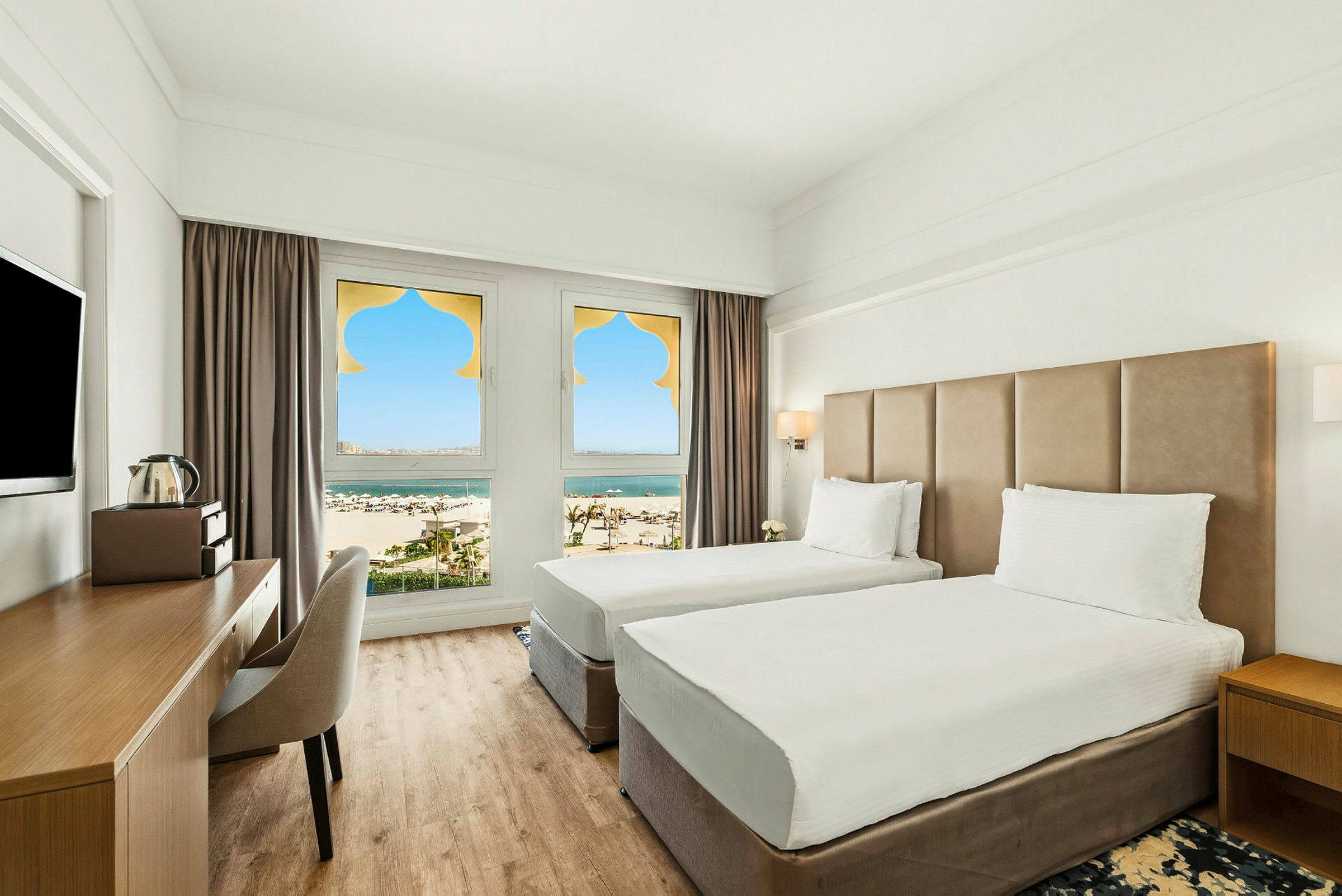 Ett hotellrum med två enkelsängar, en skrivbordsplats och fönster med utsikt över en sandstrand och hav. (AI-genererad text)
