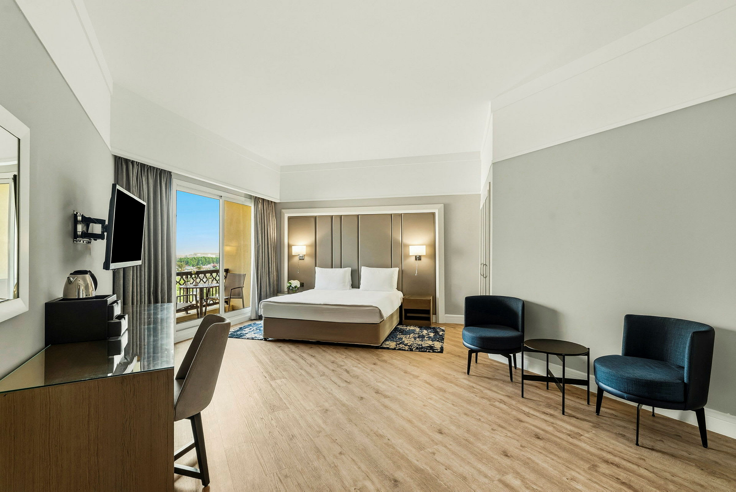 Ett modernt hotellrum med en dubbelsäng, en sittgrupp med stolar och bord, samt en arbetsyta. Utgång till balkong synlig genom stora fönster. (AI-genererad text)