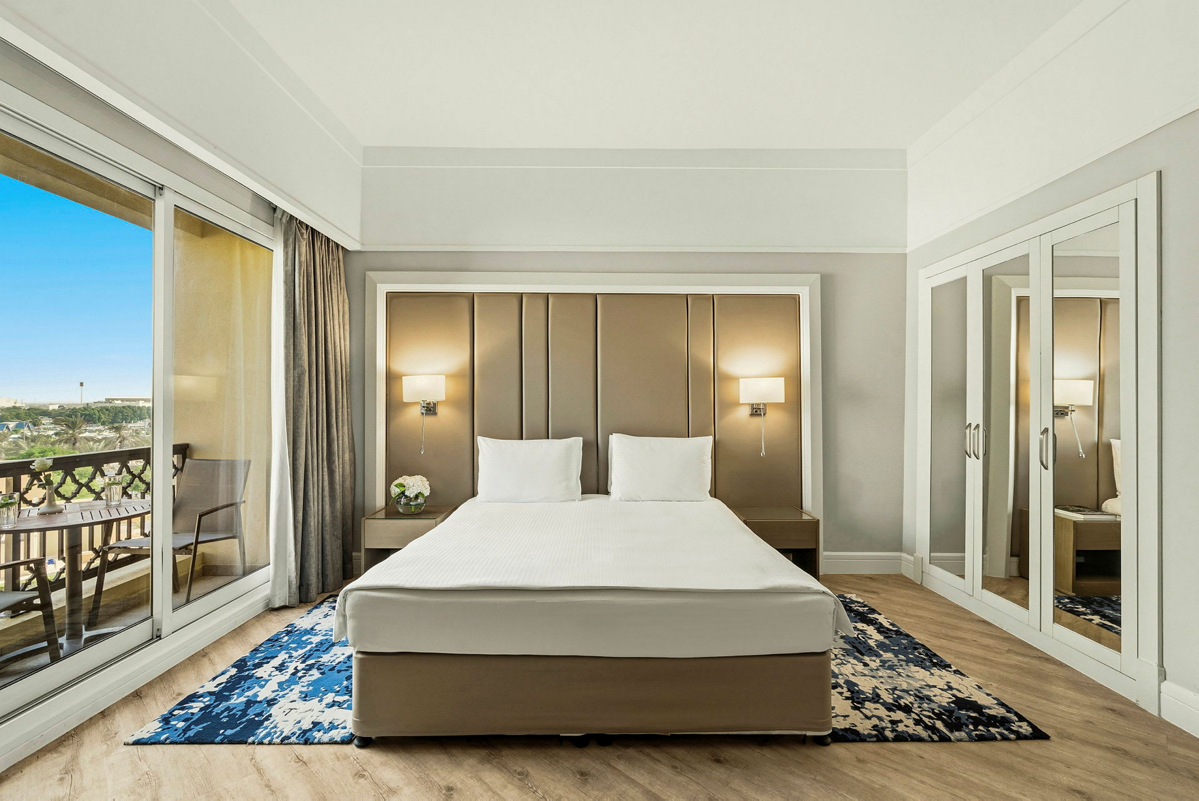 Ett hotellrum med en dubbelsäng, två sänglampor, en matta och balkong med glasdörr. (AI-genererad text)
