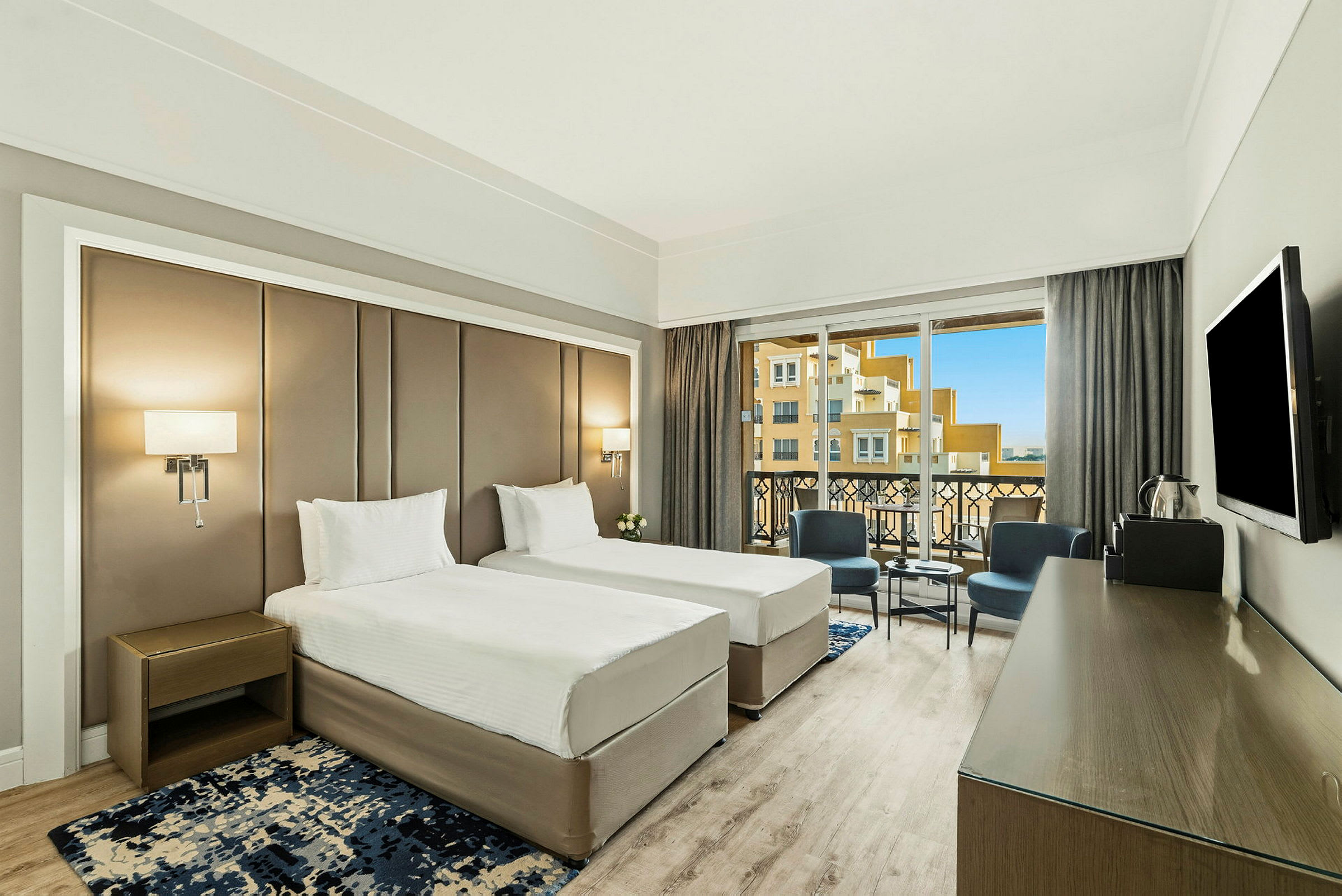 Ett hotellrum med två enkelsängar, en balkong med utsikt och möbler inklusive stolar och bord. (AI-genererad text)