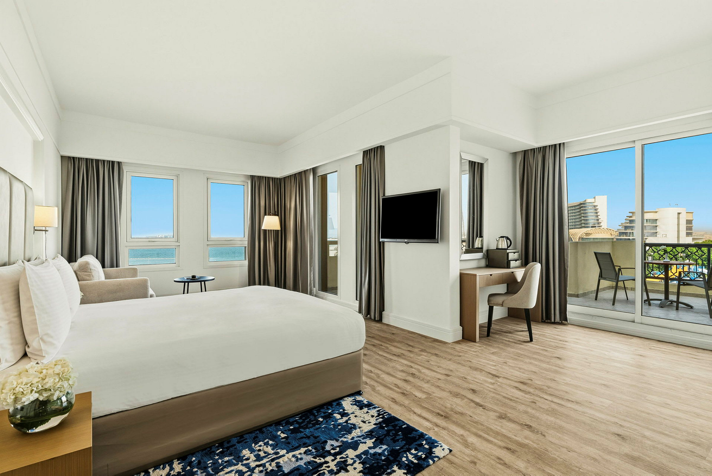 Ett modernt hotellrum med en stor säng, ett skrivbord, balkong och utsikt över en blå himmel och byggnader. (AI-genererad text)