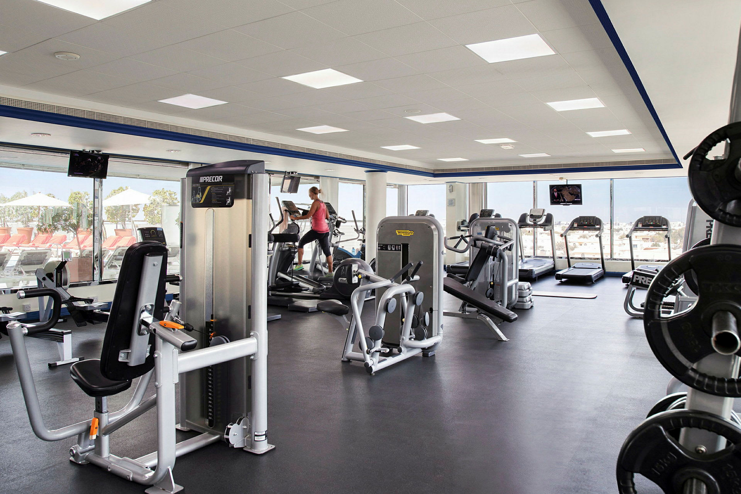 Ett gym med träningsmaskiner, vikter och ett löpband intill stora fönster som vetter mot en utomhusterrass. (AI-genererad text)