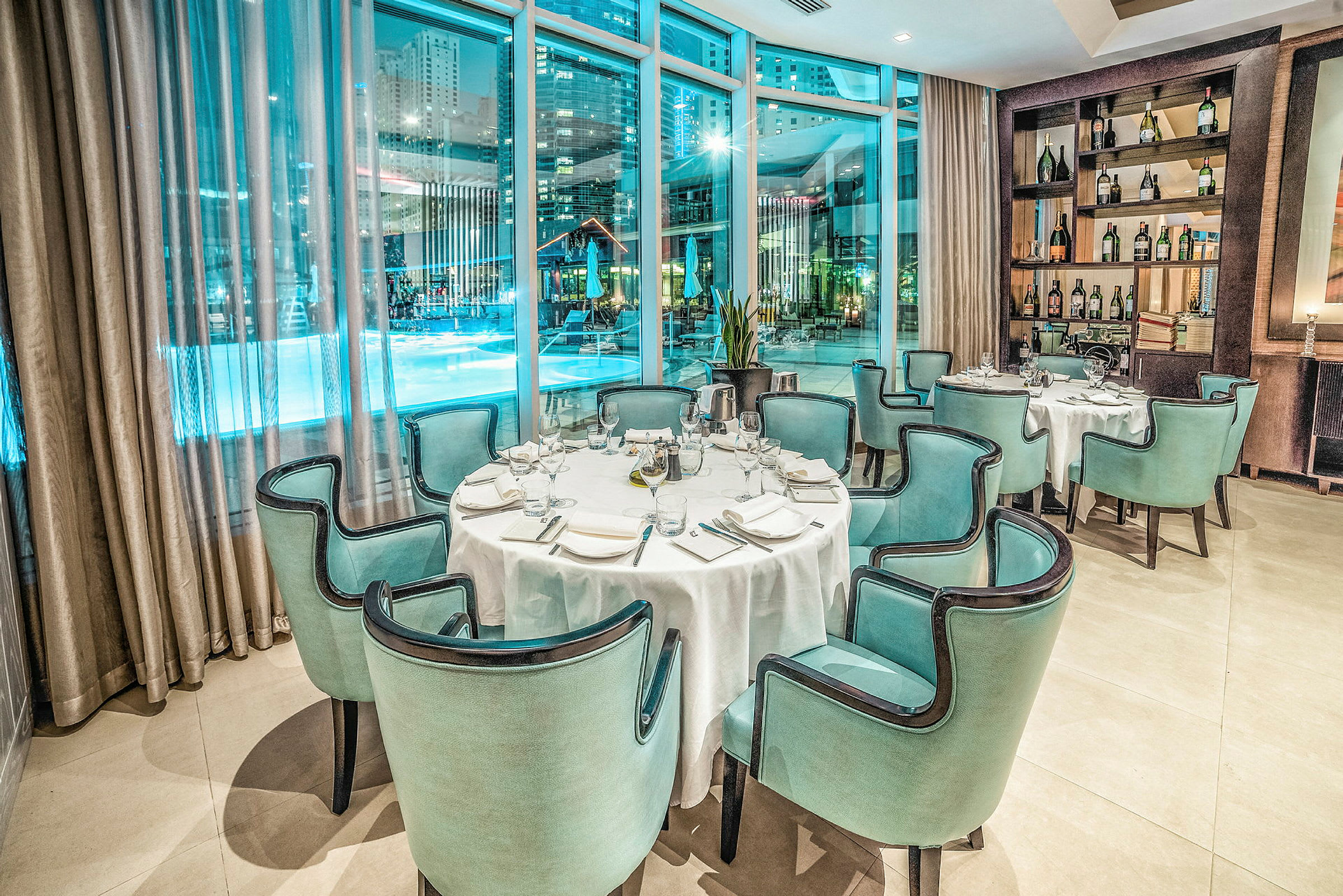 En elegant restaurang med dukade bord och grönblå stolar, belägen vid stora fönster som erbjuder utsikt över ett upplyst poolområde på natten. (AI-genererad text)