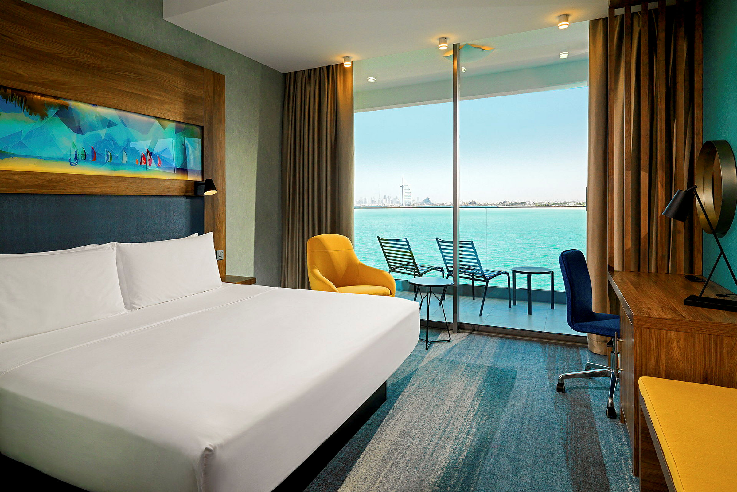 Bilden visar ett modernt hotellrum med en stor dubbelsäng, en gul fåtölj, ett skrivbord och en balkong med utsikt över havet och stadens skyline. (AI-genererad text)