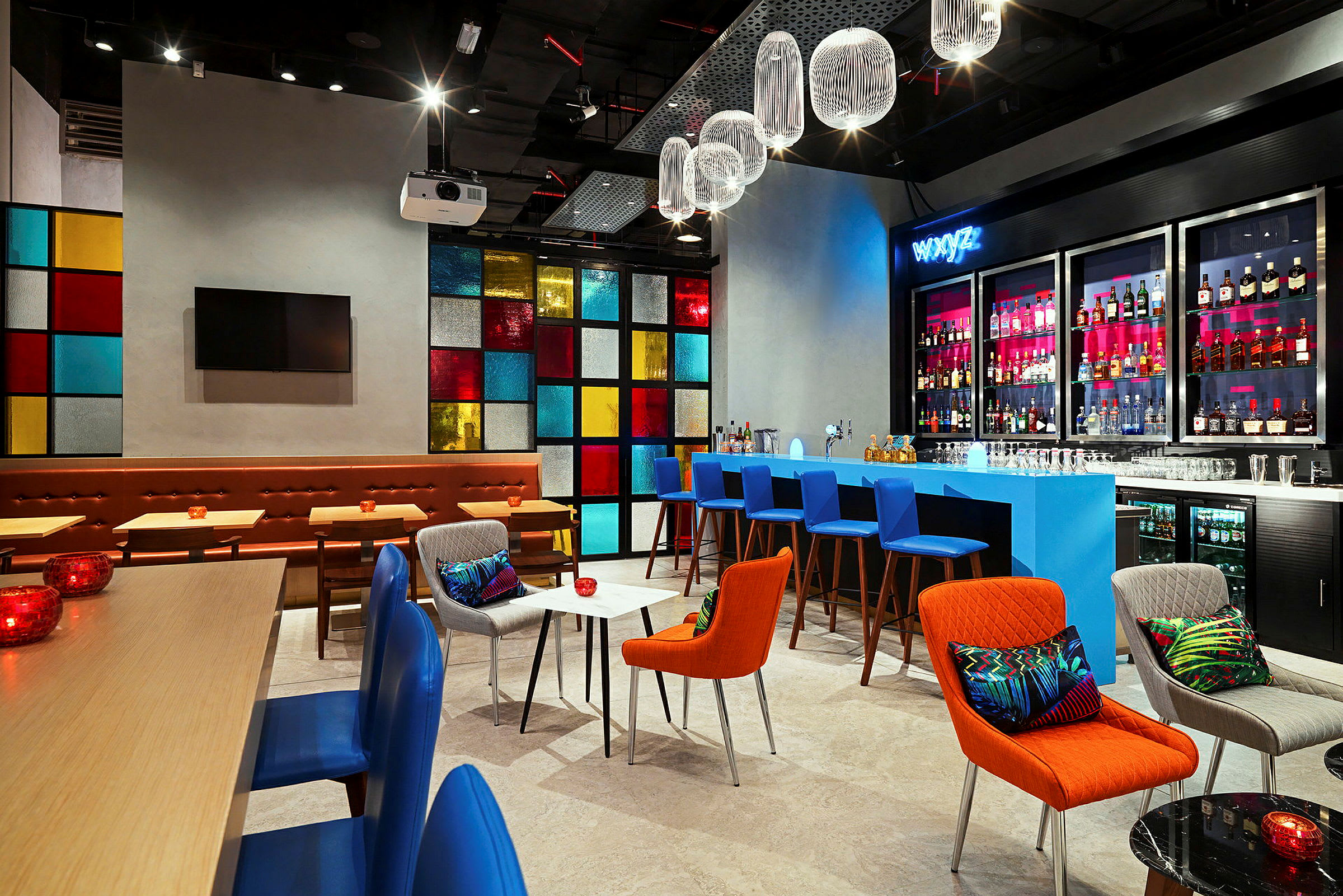 En modernt inredd bar eller lounge med färgglada möbler, ett blått bardiskområde och hyllor med flaskor bakre. Ett fönsterpanel med olika färgade rutor ger ett levande bakgrund. Belysningen är varm och designen är elegant och stilren. (AI-genererad text)