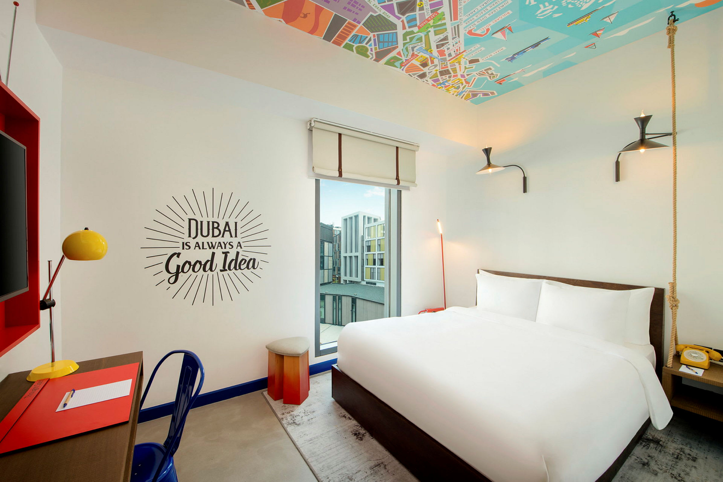 Ett hotellrum med en vit dubbelsäng, skrivbord med lampa och en väggtext som lyder 'Dubai is always a good idea'. (AI-genererad text)