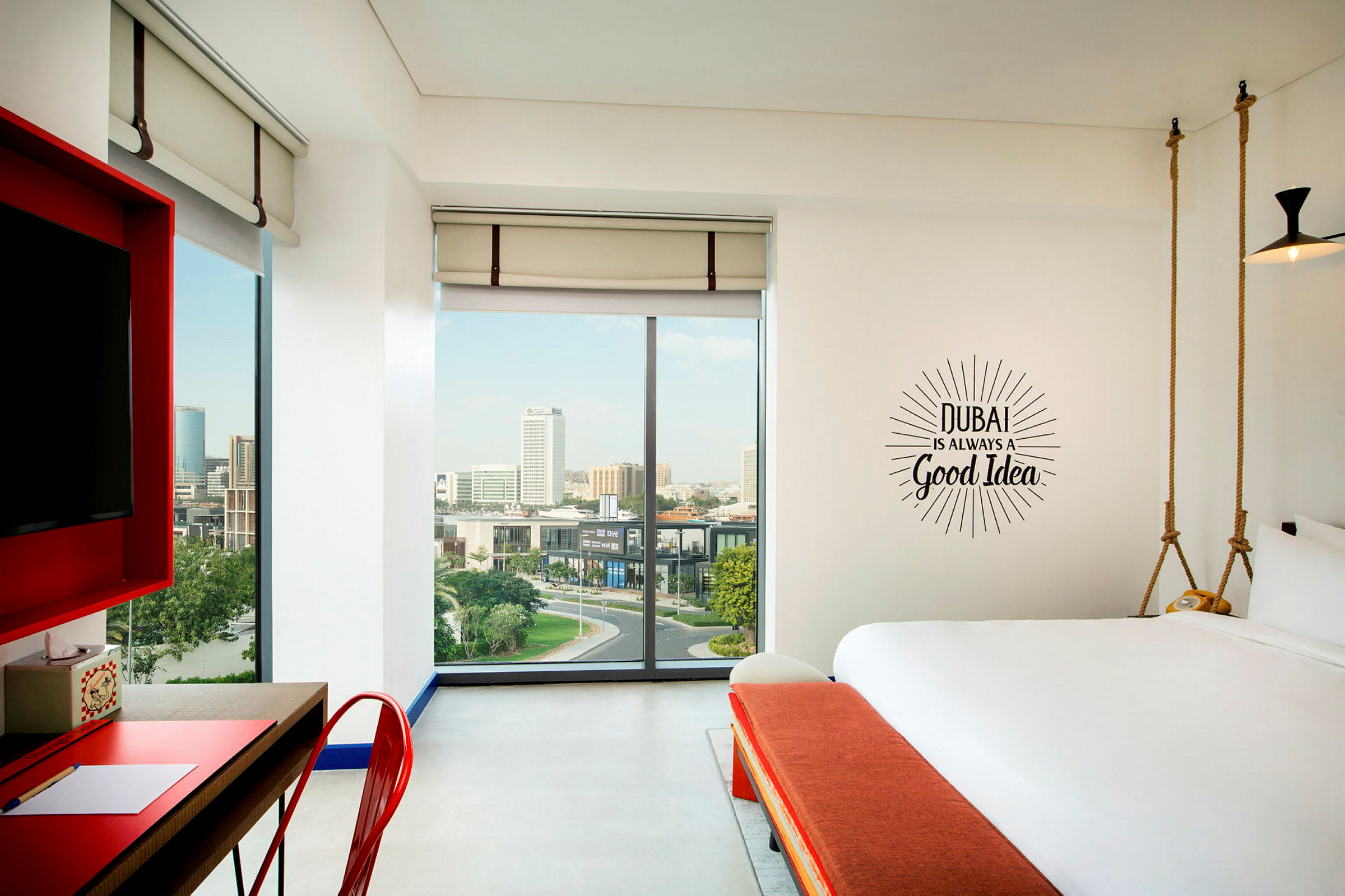 Ett modernt hotellrum med stora fönster och utsikt över en stad. På väggen står texten: "Dubai is always a good idea". (AI-genererad text)