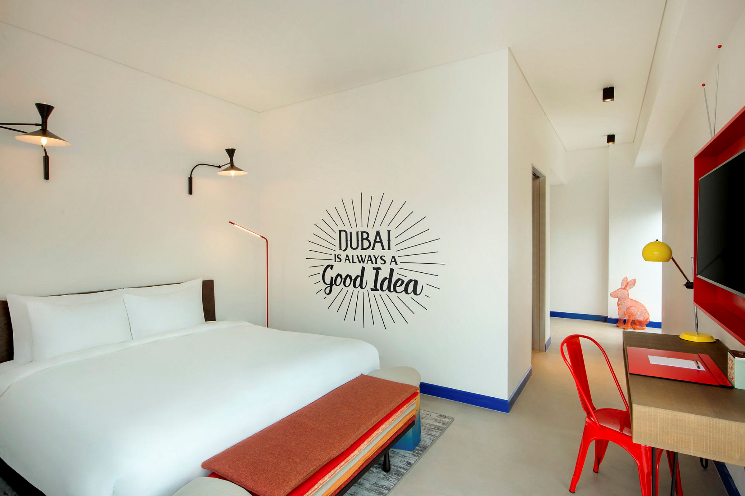 Ett hotellrum med en dubbelsäng, en väggtext som lyder "Dubai is always a good idea", och inredning i moderna färger. (AI-genererad text)