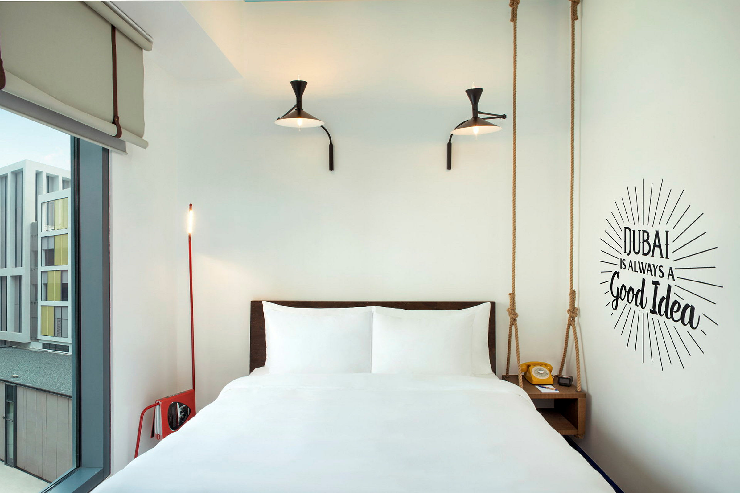 Ett modernt hotellrum med en säng, två lampor på väggen och en väggtext som lyder "Dubai is always a good idea". (AI-genererad text)