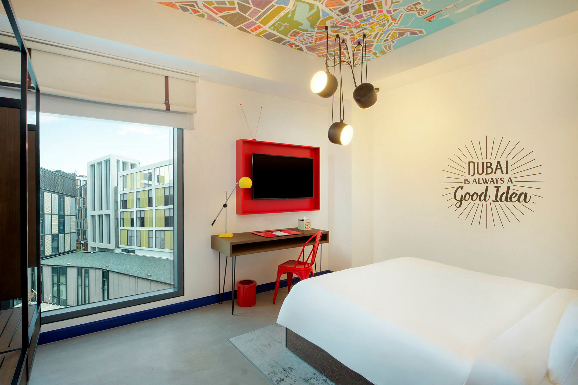Ett ljust hotellrum med en stor säng, skrivbord, och en vägg-TV. Fönstret visar byggnader utanför. (AI-genererad text)