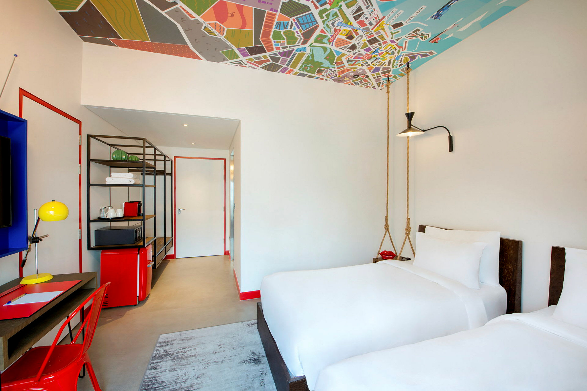 Ett modernt hotellrum med två vita enkelsängar, ett skrivbord, en röd stol och färgglada inredningsdetaljer. Taket är dekorerat med ett kartmönster. (AI-genererad text)