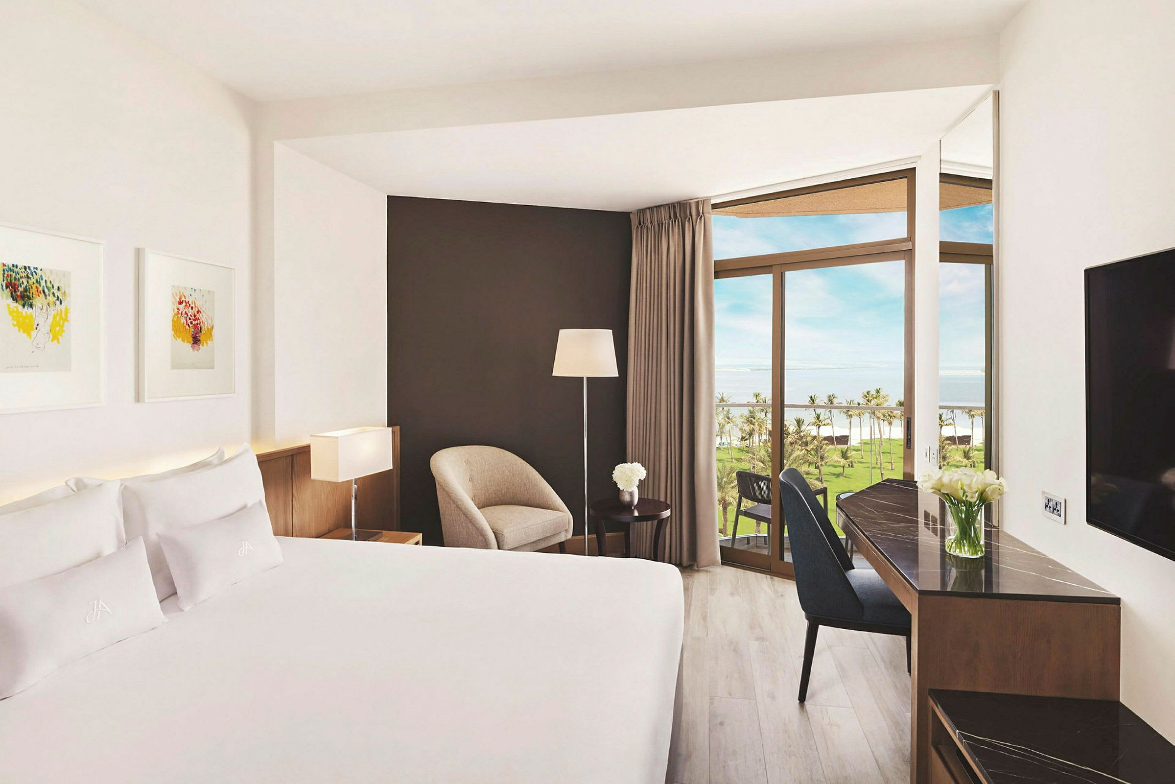 Ett hotellrum med en stor säng, ett skrivbord, en stol och stora fönster som vetter mot en balkong med utsikt över en gräsyta och palmer. (AI-genererad text)