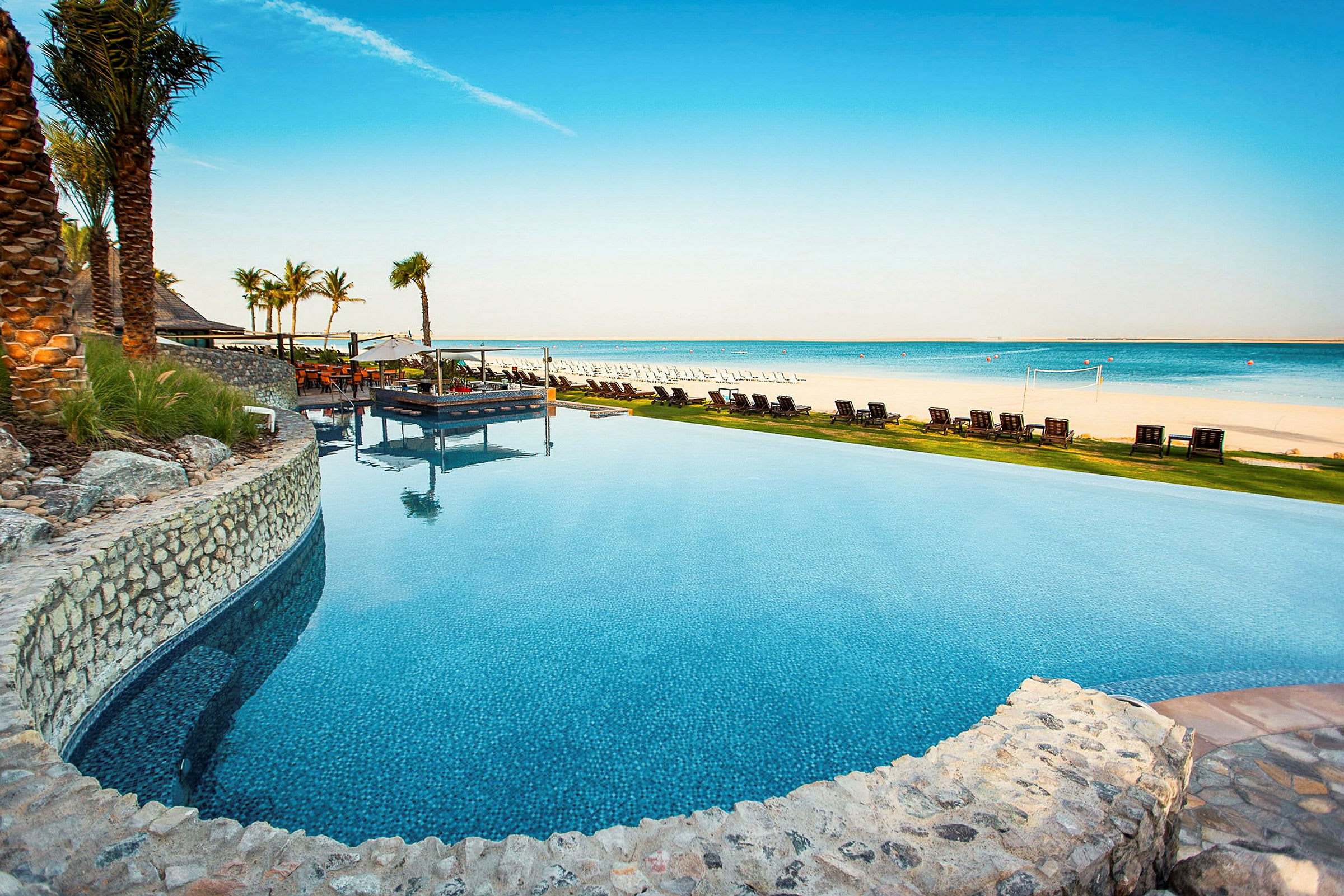 Bild som visar en infinity-pool vid en anläggning vid havet med sandstrand och solsängar i bakgrunden. (AI-genererad text)