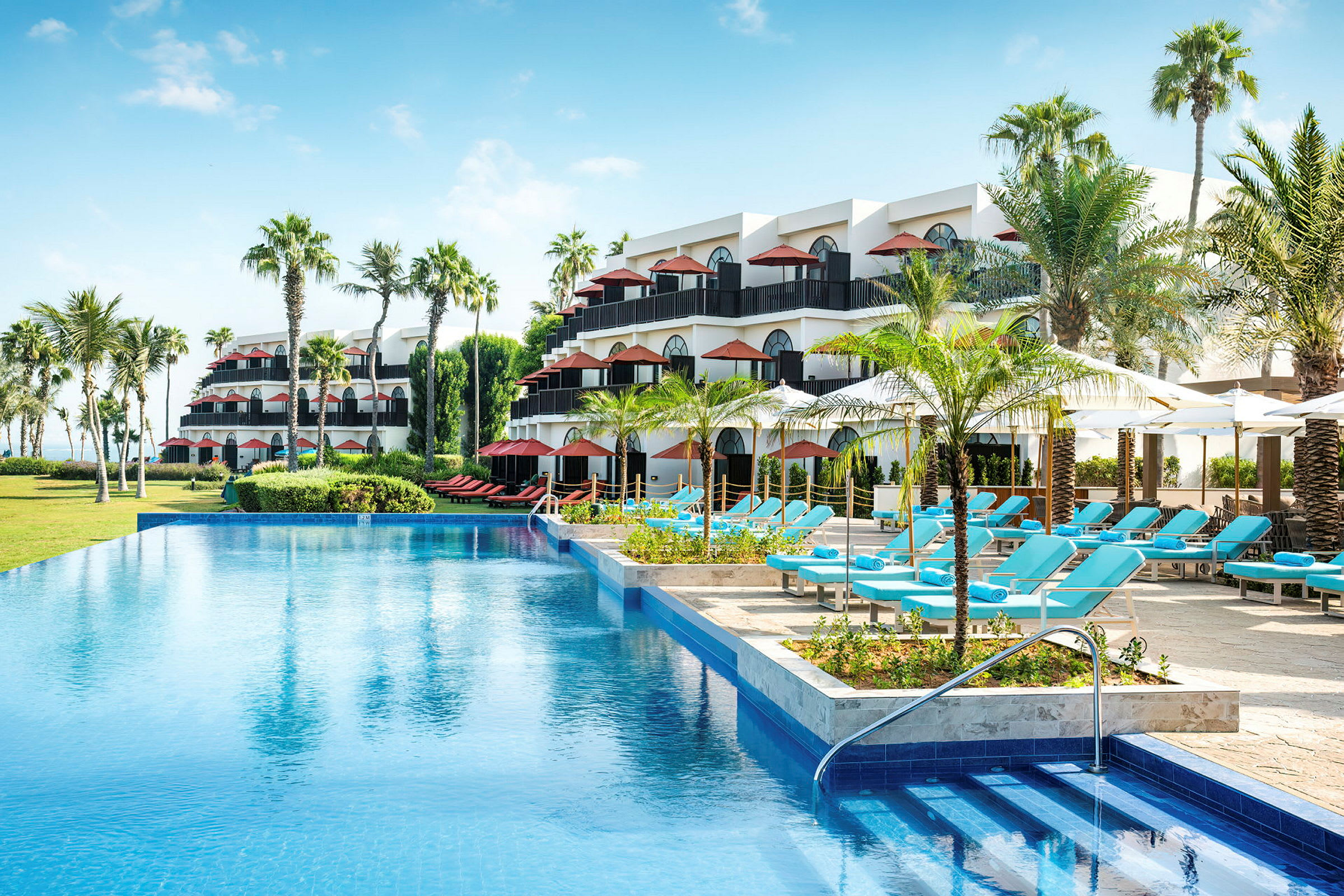 Ett hotellområde med flera våningar, palmer, en swimmingpool och solstolar under parasoll. (AI-genererad text)