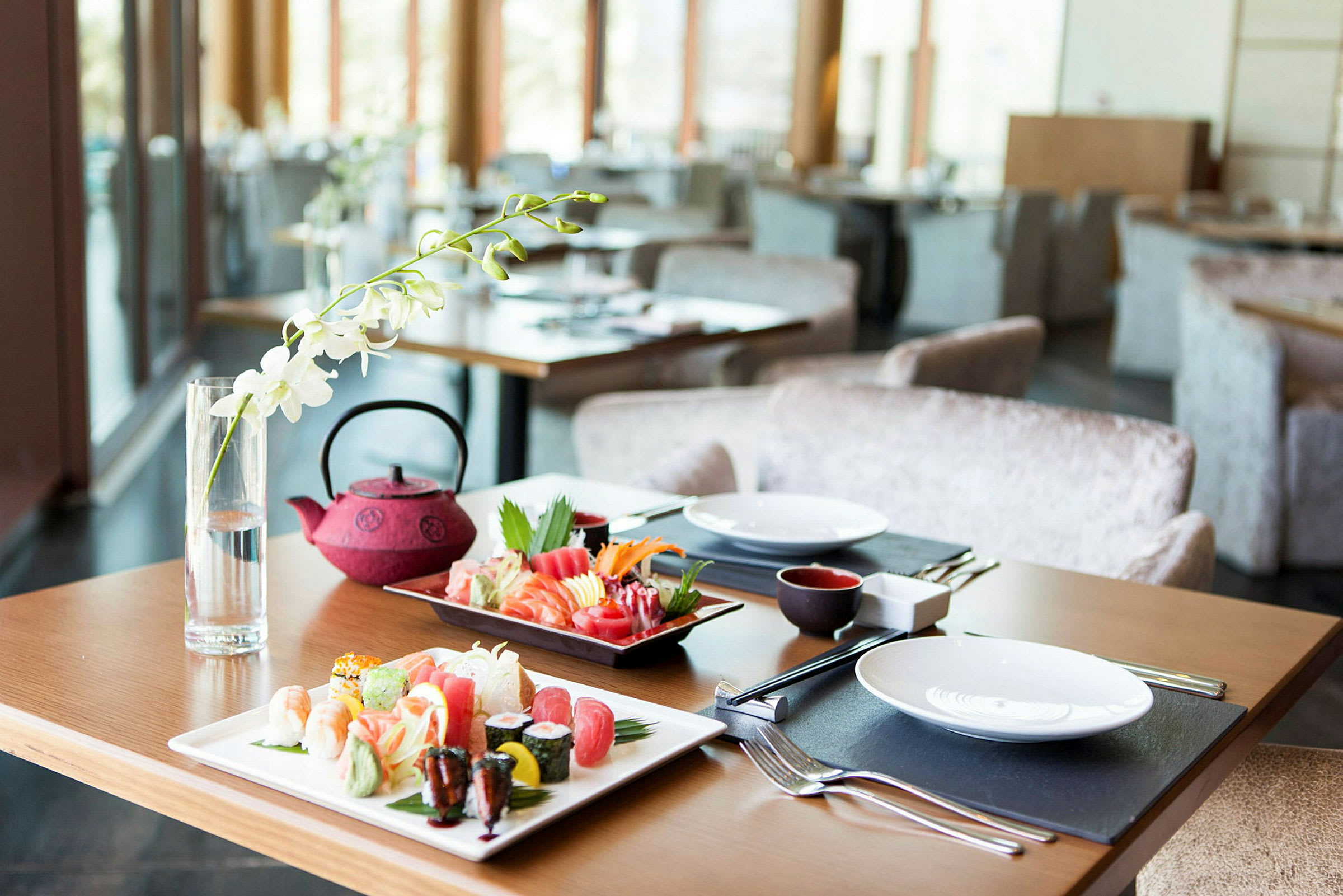 Bild av ett dukat bord i en restaurang med sushi och en röd tekanna. Dekorationer och stora fönster syns i bakgrunden. (AI-genererad text)
