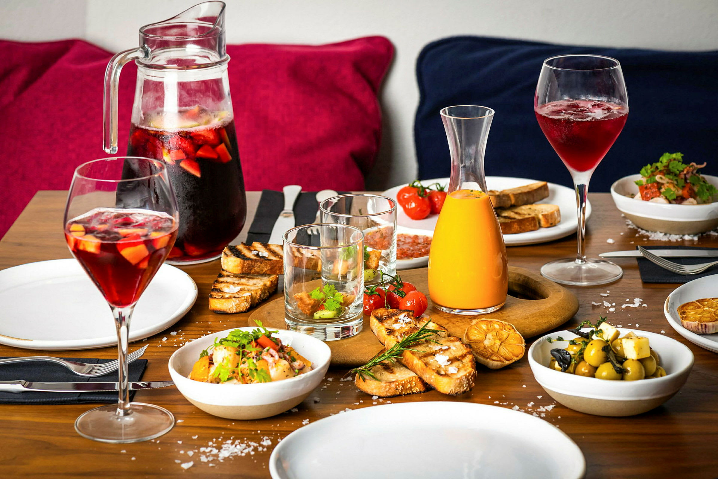 Ett dukat bord med olika maträtter och drycker, inklusive bröd, sallad, oliver och karaffer med juice och röda drycker. (AI-genererad text)