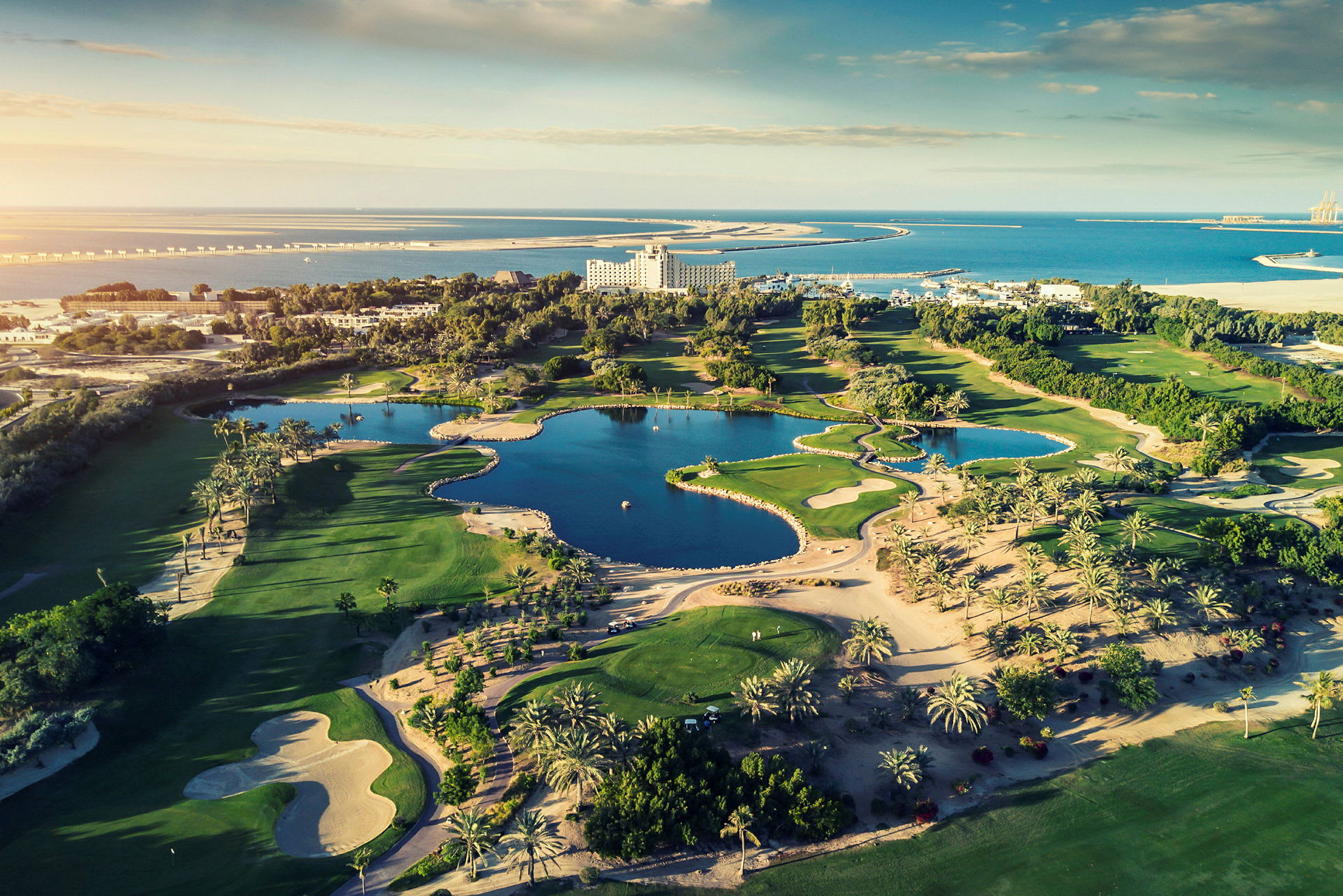 Flygfoto över en golfbana med vattenhinder och omgivande vegetation, samt hav och byggnader i bakgrunden. (AI-genererad text)