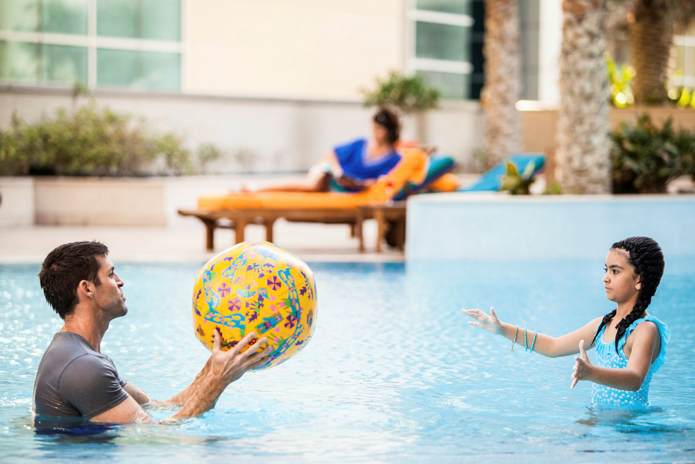 Två personer leker i en pool med en färgglad strandboll. I bakgrunden syns en person som kopplar av på en solstol. (AI-genererad text)