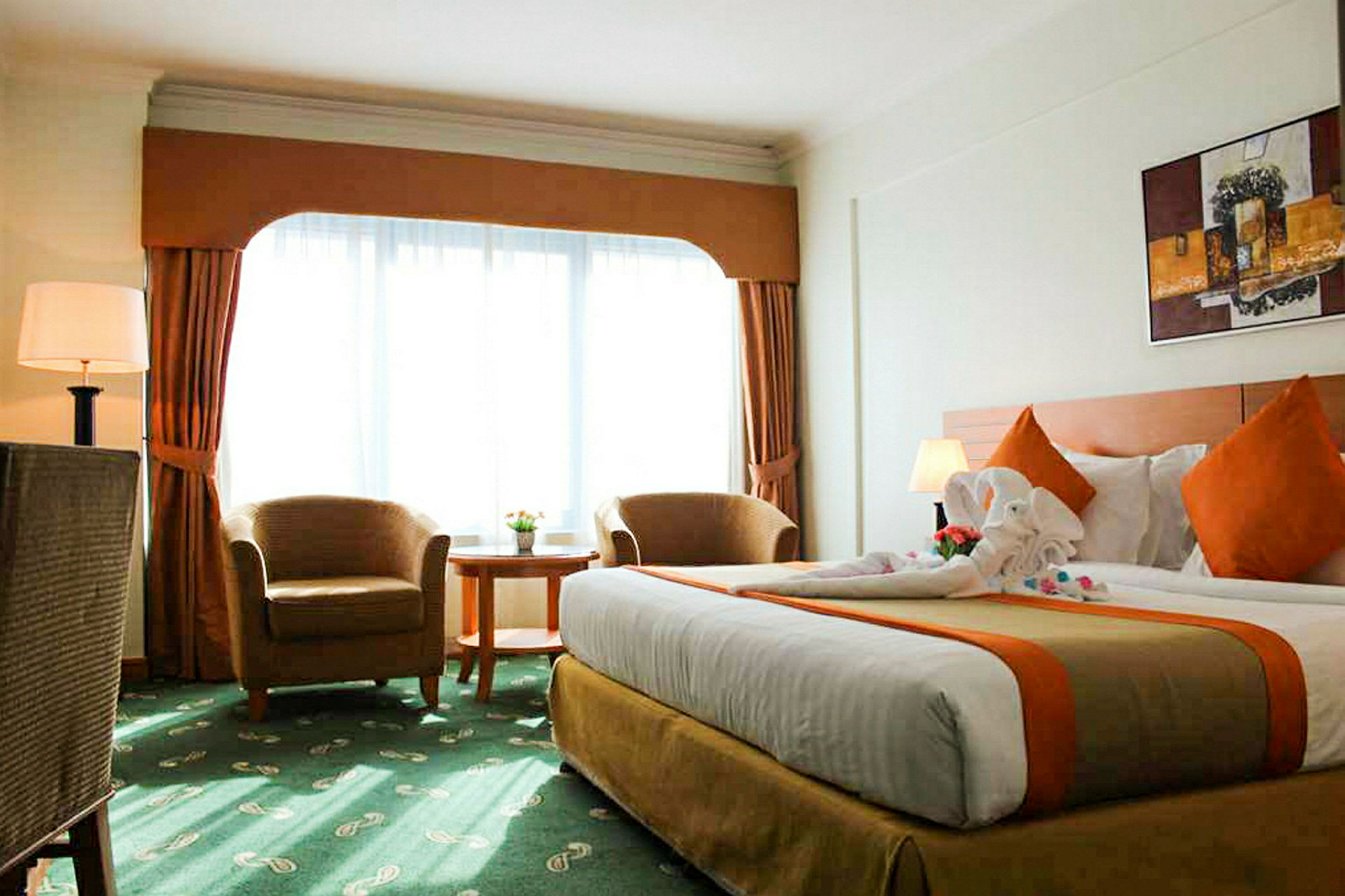 Ett hotellrum med en stor säng, två fåtöljer, ett runt bord och en stor fönsteryta med gardiner. (AI-genererad text)