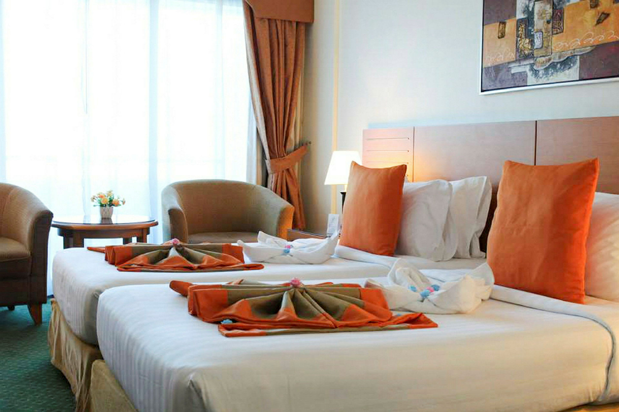 Ett hotellrum med två sängar bäddade med vita lakan och orange kuddar. Det finns en sittgrupp med två fåtöljer och ett litet bord framför ett stort fönster, samt en tavla på väggen ovanför sängarna. (AI-genererad text)