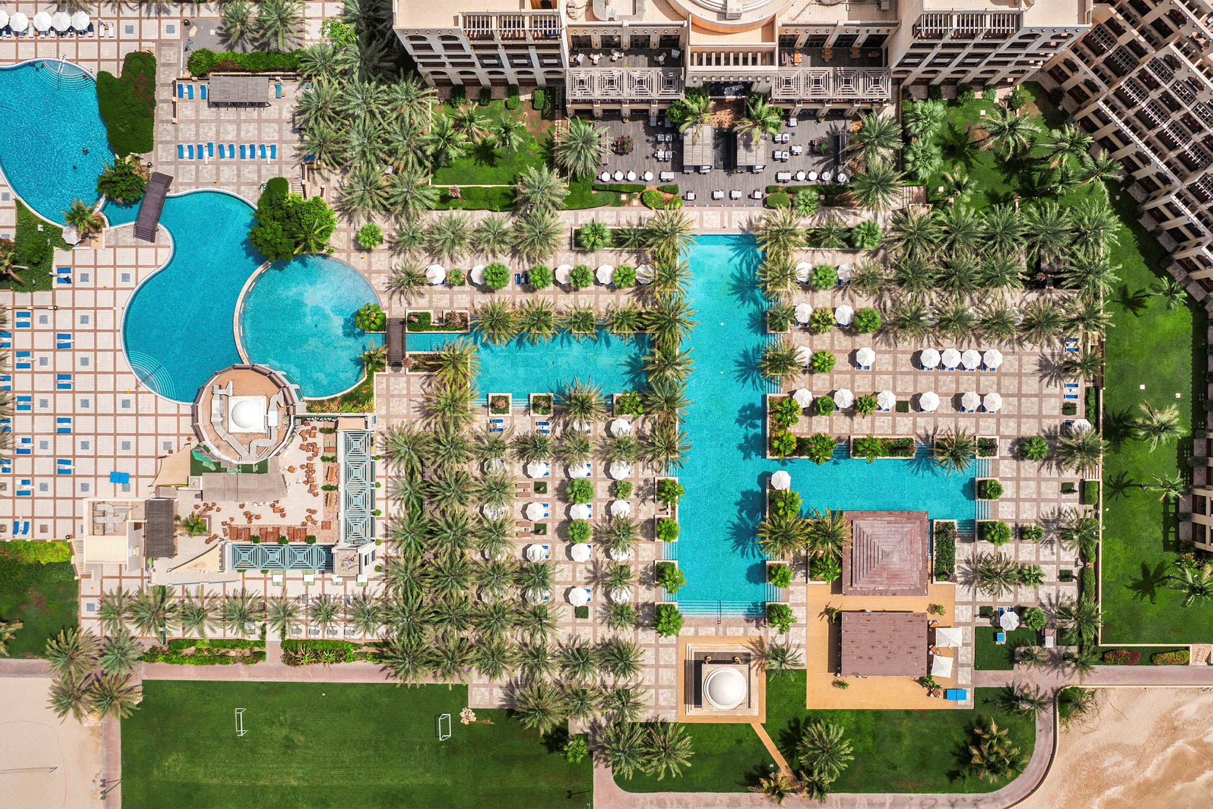 Flygfoto över poolområdet på en resort med flera bassänger, parasoller och träd. (AI-genererad text)