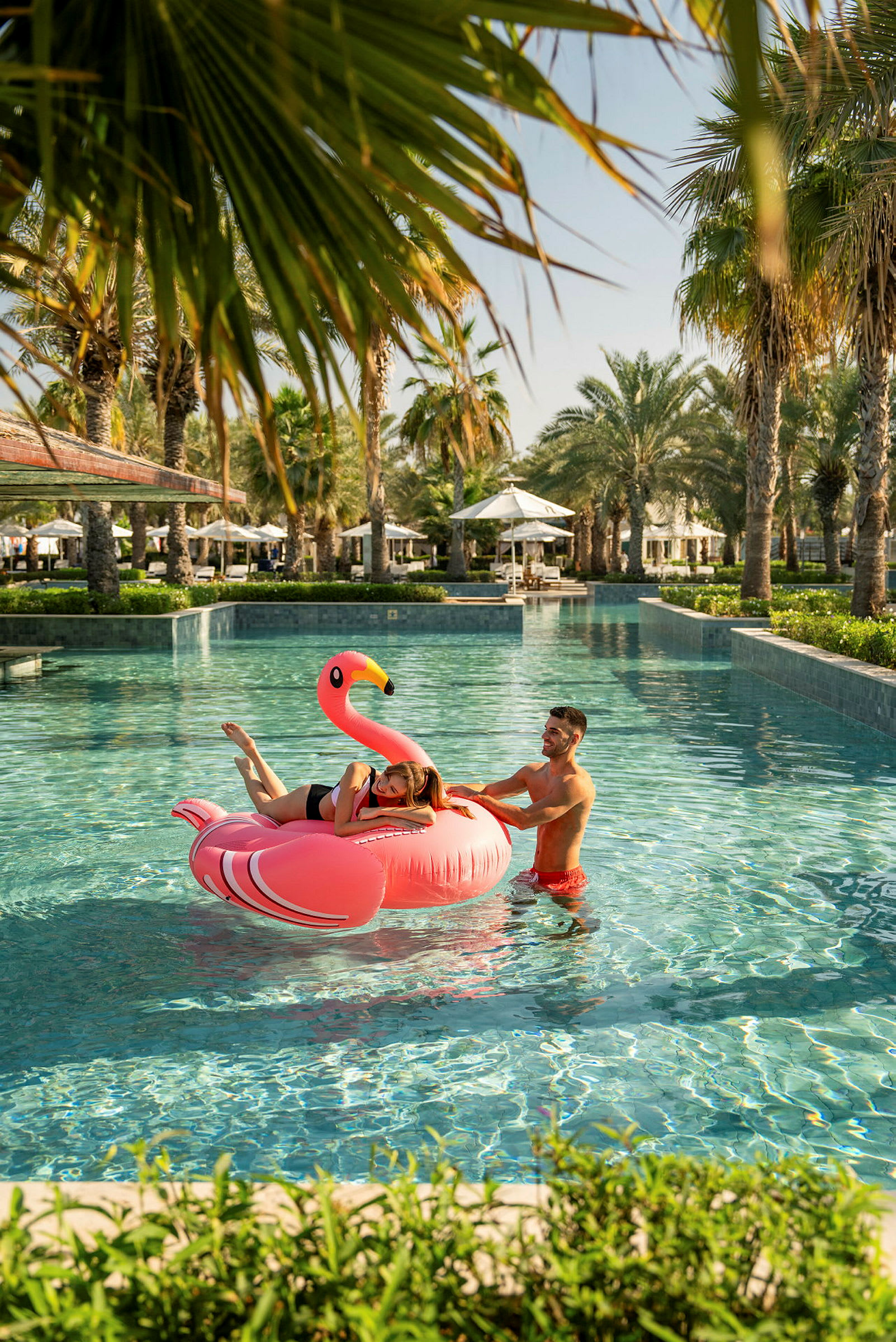 Två personer vid en pool med en uppblåsbar flamingo. Palmer och solstolar syns i bakgrunden. (AI-genererad text)