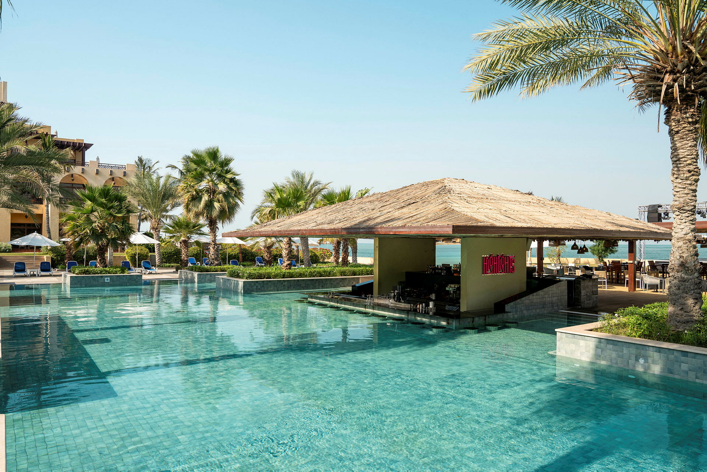 En pool med klart vatten och en poolbar under ett halmtak, omgiven av solstolar och palmer. (AI-genererad text)
